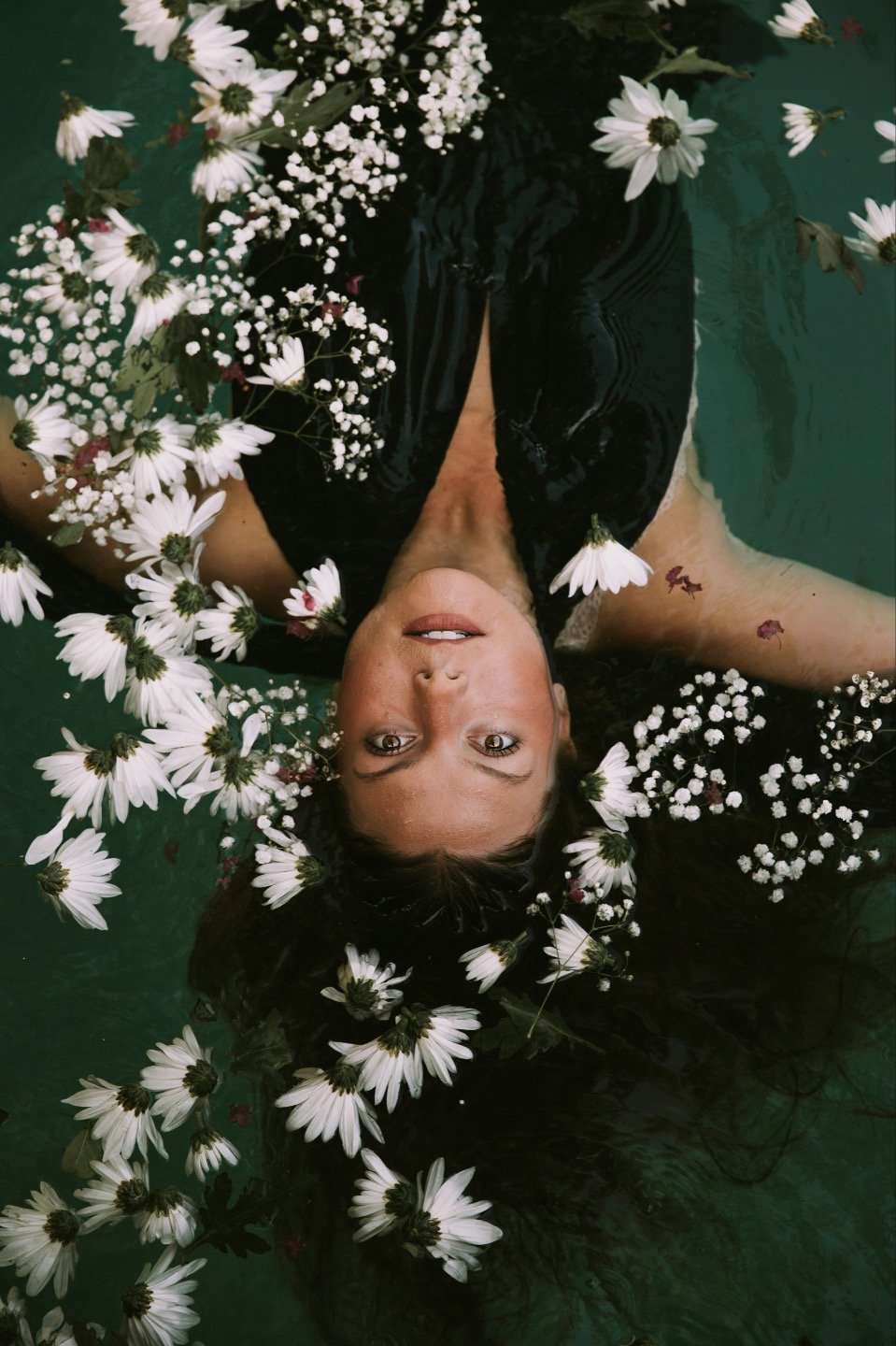A woman lying on her back in a green body of water, surrounded by white flowers and baby's breath, with her head submerged and her face looking up at the camera.