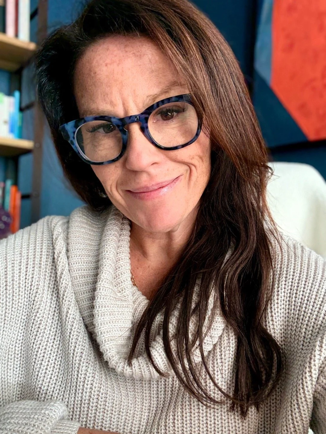 A woman with brown hair and glasses wearing a beige knit sweater taking a selfie indoors.