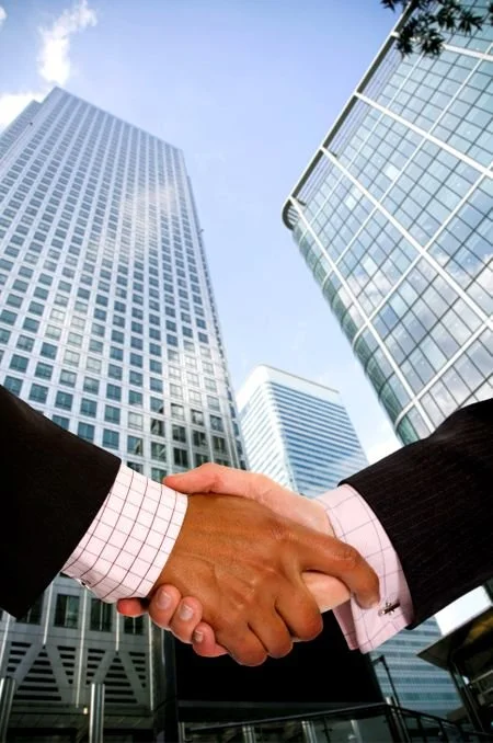 Two people in business suits shaking hands in front of tall skyscrapers in a city.