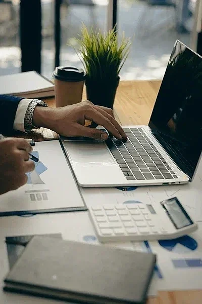 Person working at a desk with a laptop, paper documents, a calculator, a notebook, a coffee cup, and a potted plant.