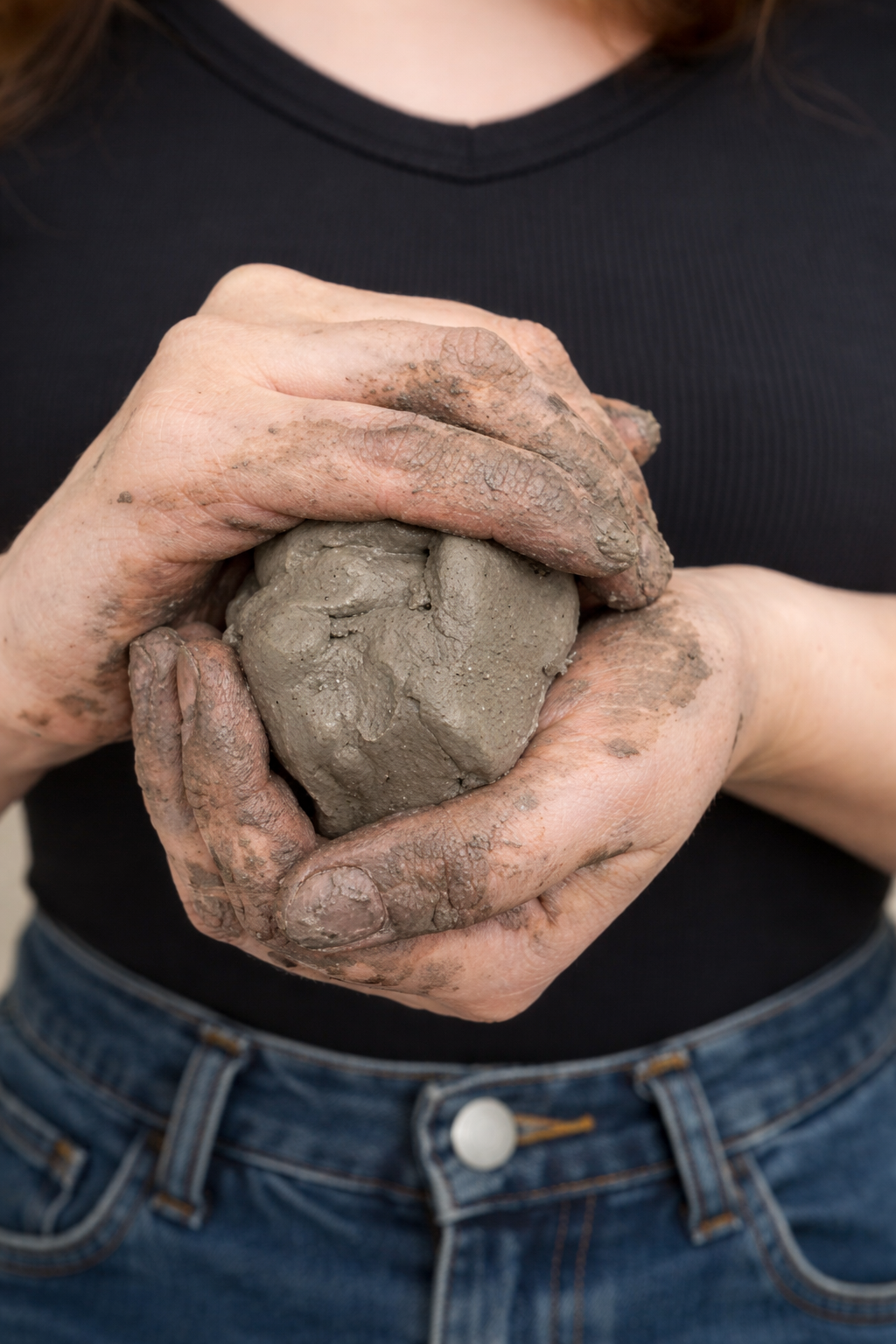 Person with muddy hands holding a ball of wet clay, wearing a black shirt and jeans.