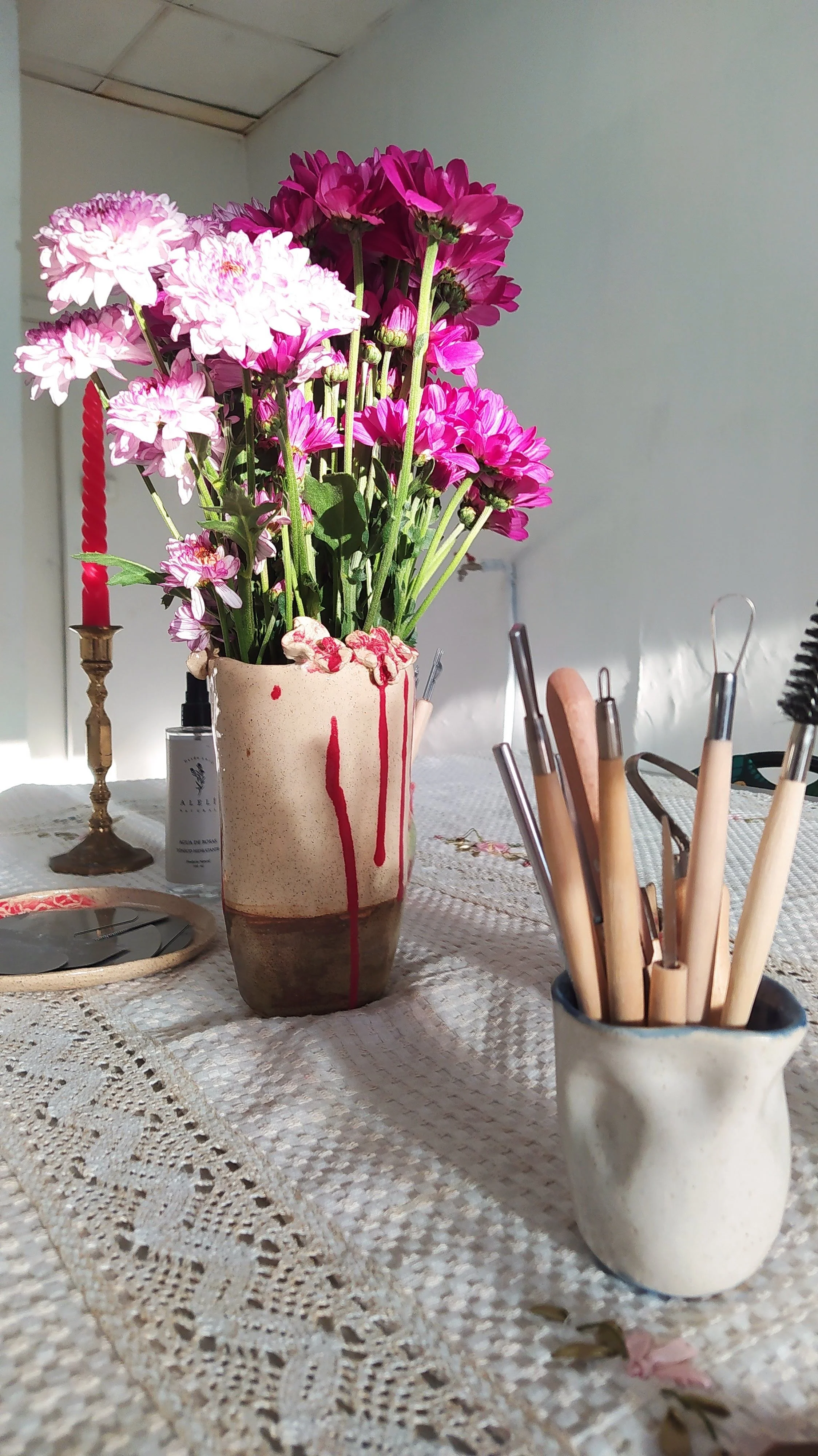 A beige vase with pink and white flowers on a lace tablecloth, with a candlestick, bottle, and utensil holder nearby.