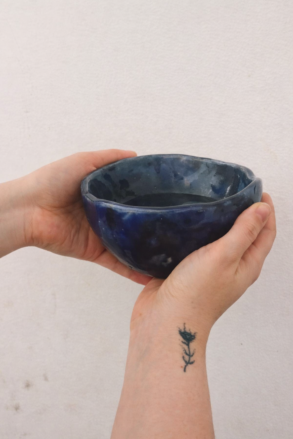 Two hands holding a dark blue handmade ceramic bowl against a plain wall, with a small tattoo of a flower on one wrist.