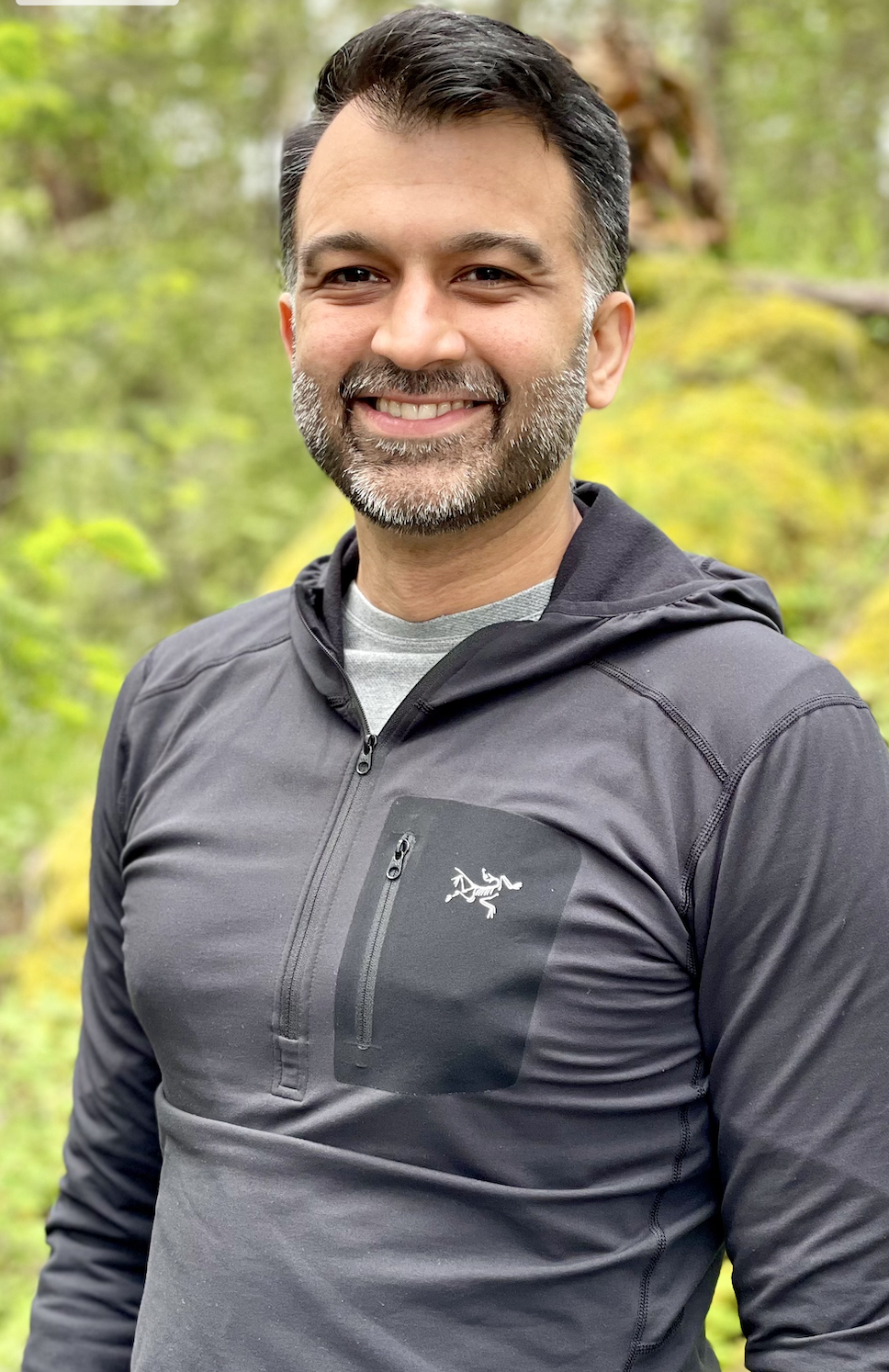Dr Bains, a smiling man with a beard and short dark hair standing outdoors in a forest setting. He is wearing a black jacket with a zippered chest pocket featuring a white scorpion logo.