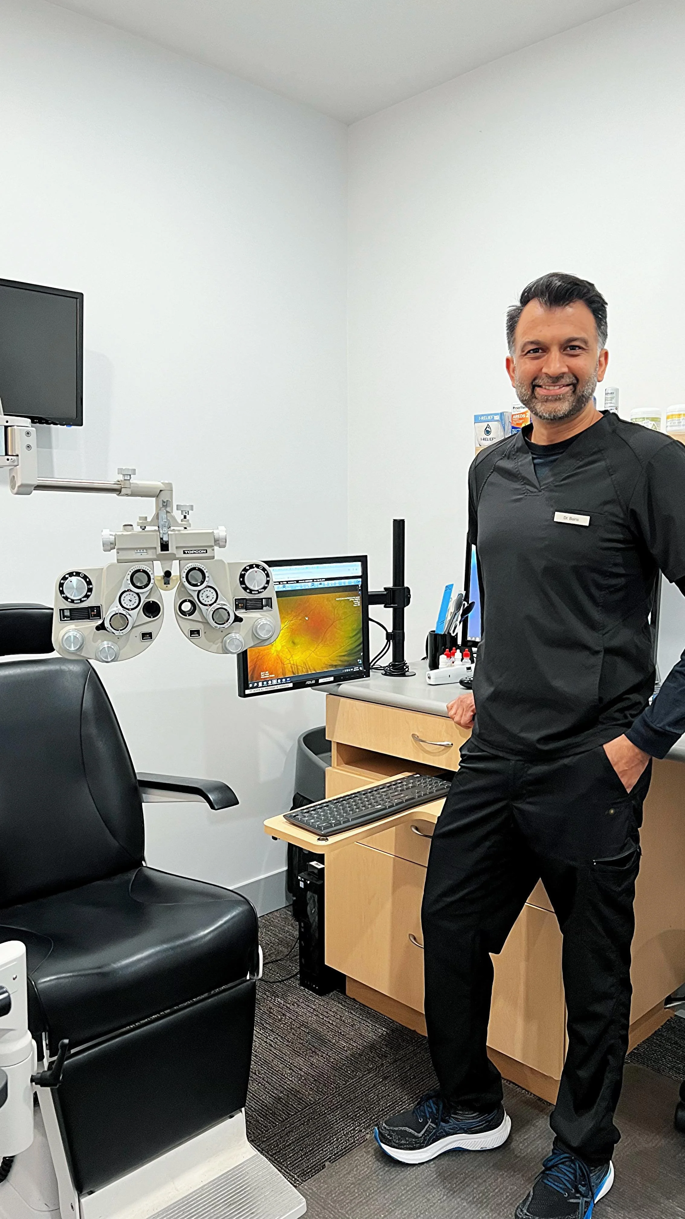 An Optometrist eye doctor standing in a medical examination room next to ophthalmology and optometry equipment including a phoropter, computer monitor displaying retina images, and other medical tools to help detect eye diseases.