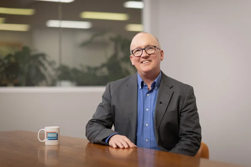 A smiling man wearing glasses and a gray suit sitting at a wooden conference table with a white mug next to him, in an office setting.