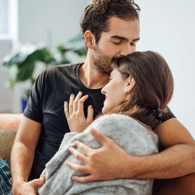 A man and woman sharing a tender moment indoors, with the woman resting her head on the man's chest and both appearing to enjoy a loving embrace.