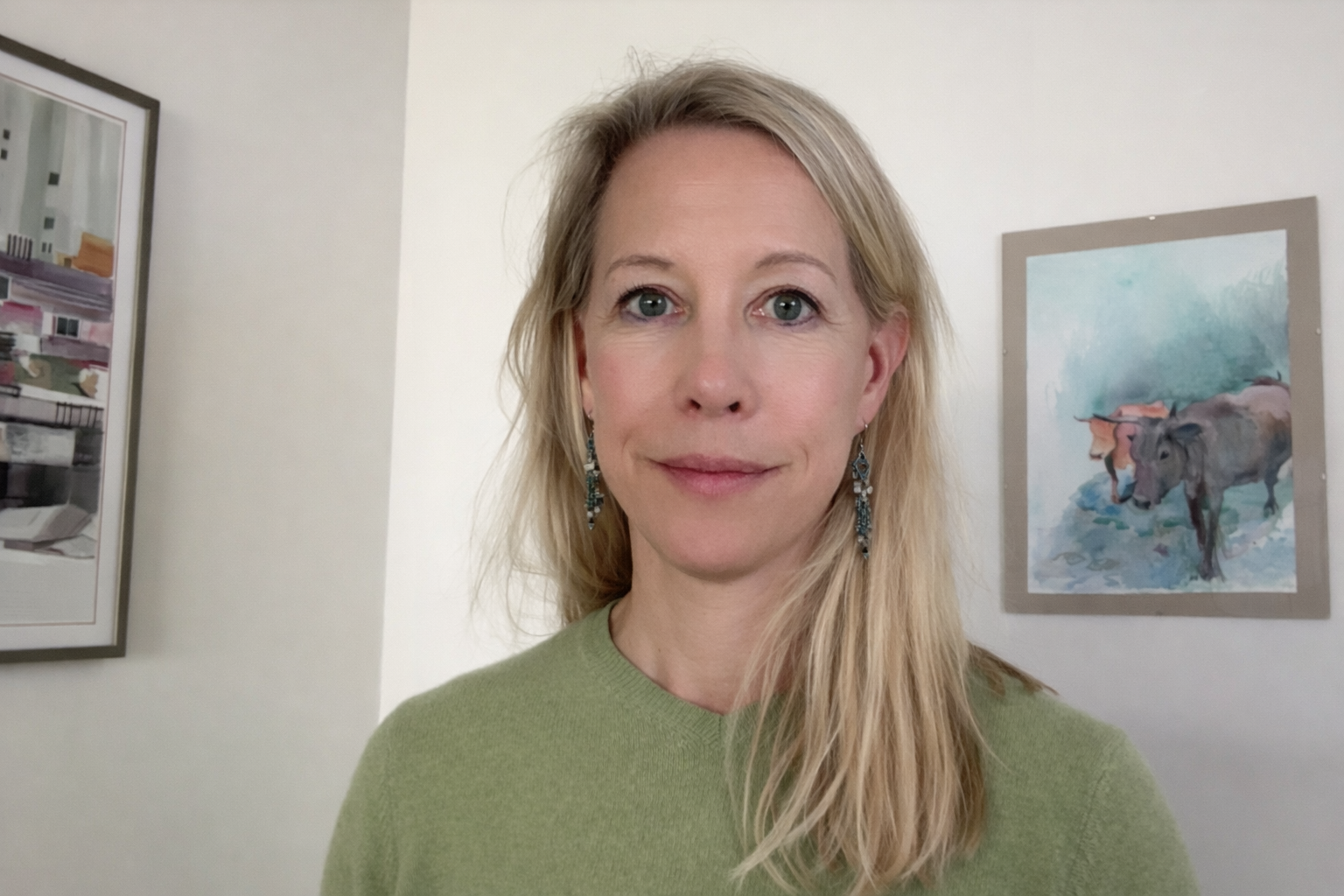A woman with long blonde hair, wearing earrings and a green top, standing in front of a white wall with framed artwork, including a colorful abstract painting and a watercolor painting of buffalo.