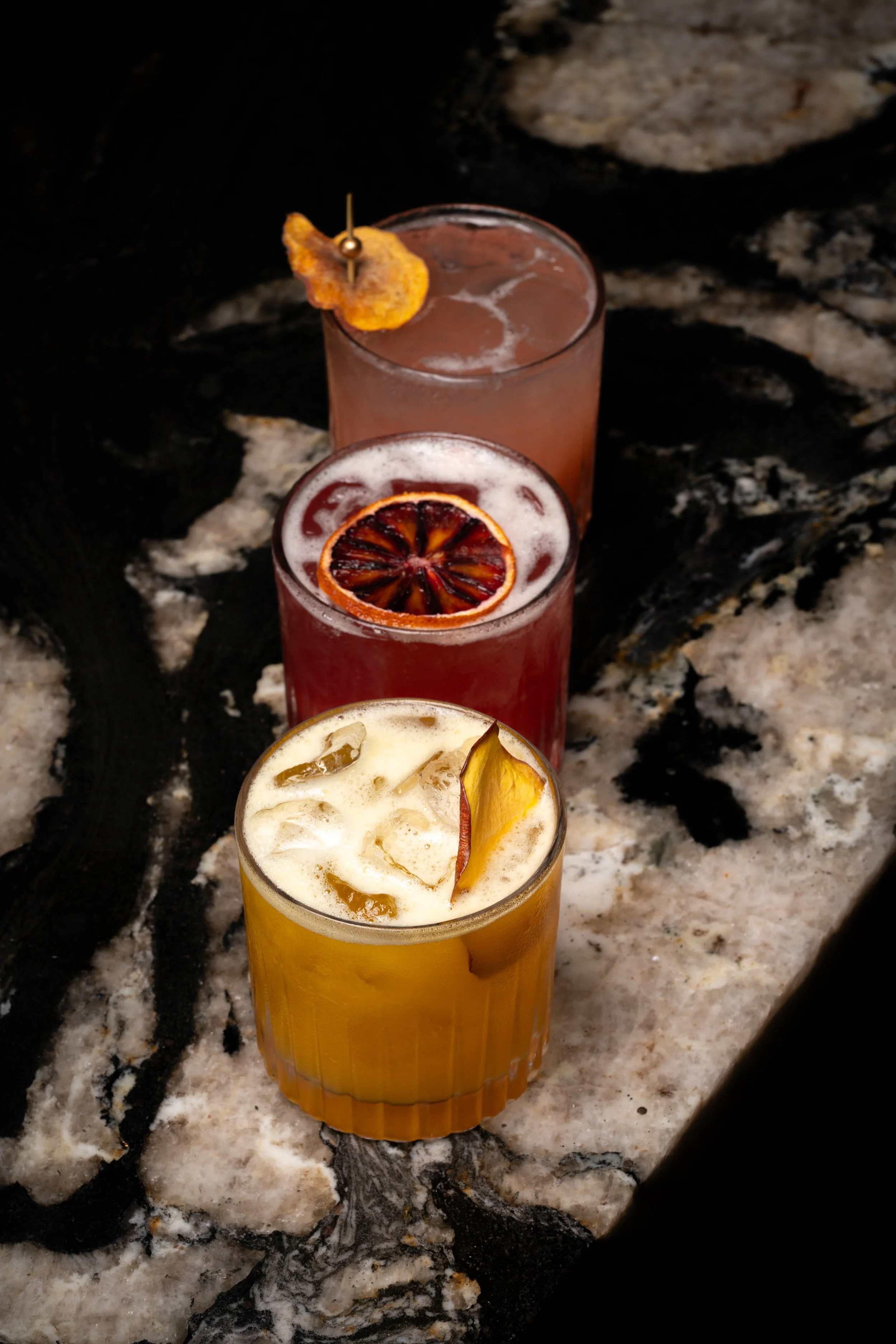 Three colorful cocktails with fruit garnishes lined up on a dark marble surface.