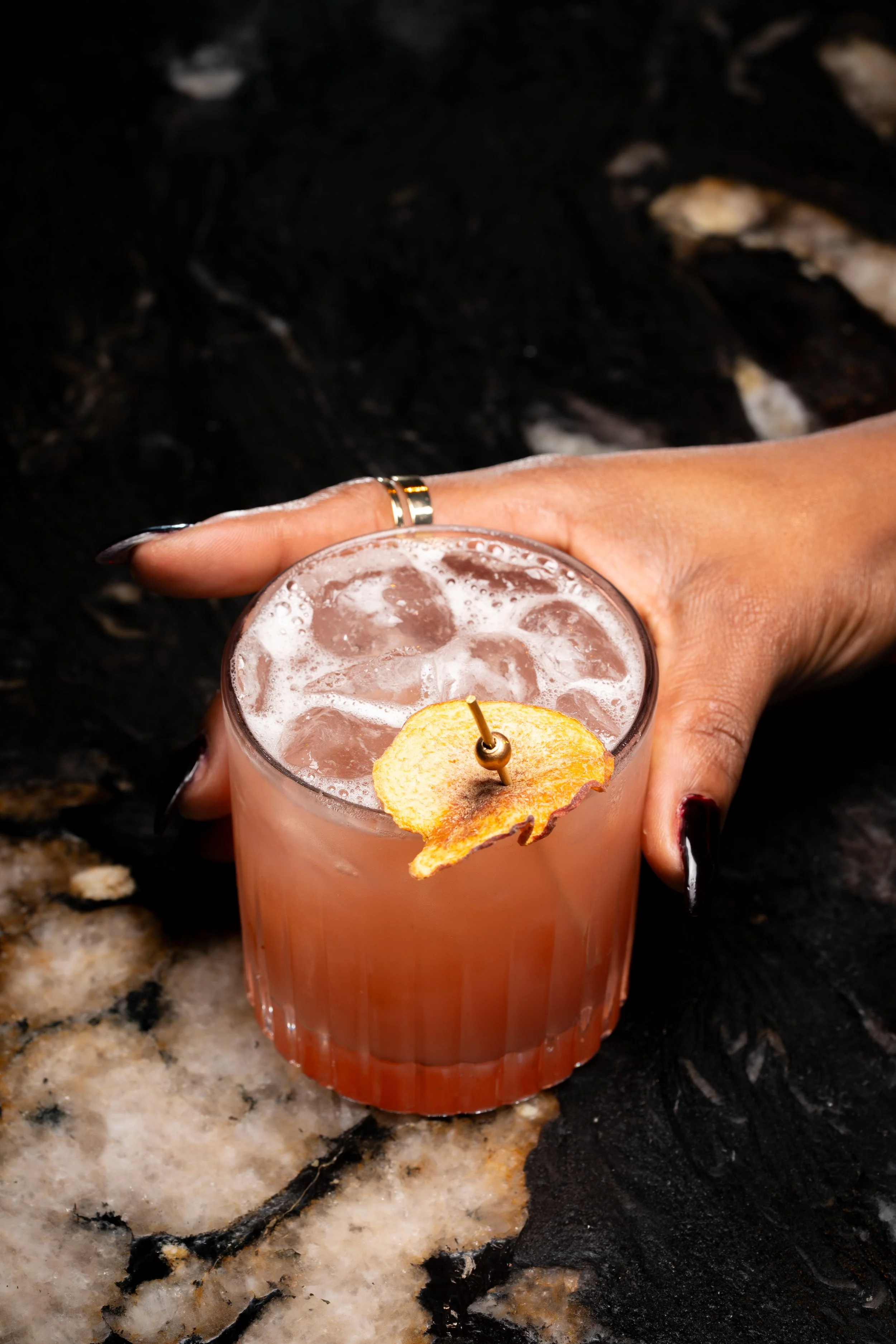 A hand holding a pink cocktail with ice on a dark marble surface.