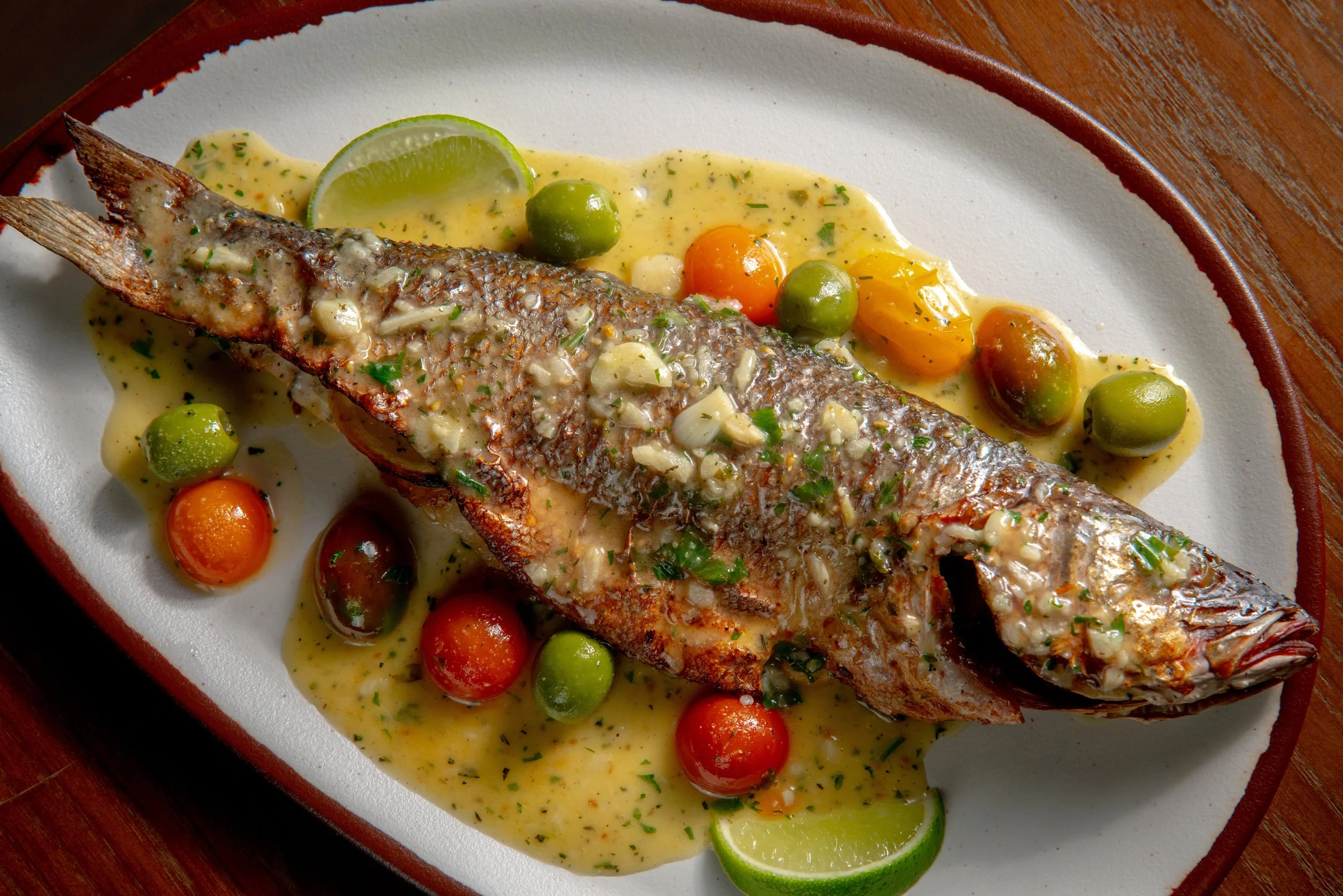 A whole cooked fish topped with garlic, herbs, and a creamy sauce, served on a plate with lime wedges, cherry tomatoes, and green olives.