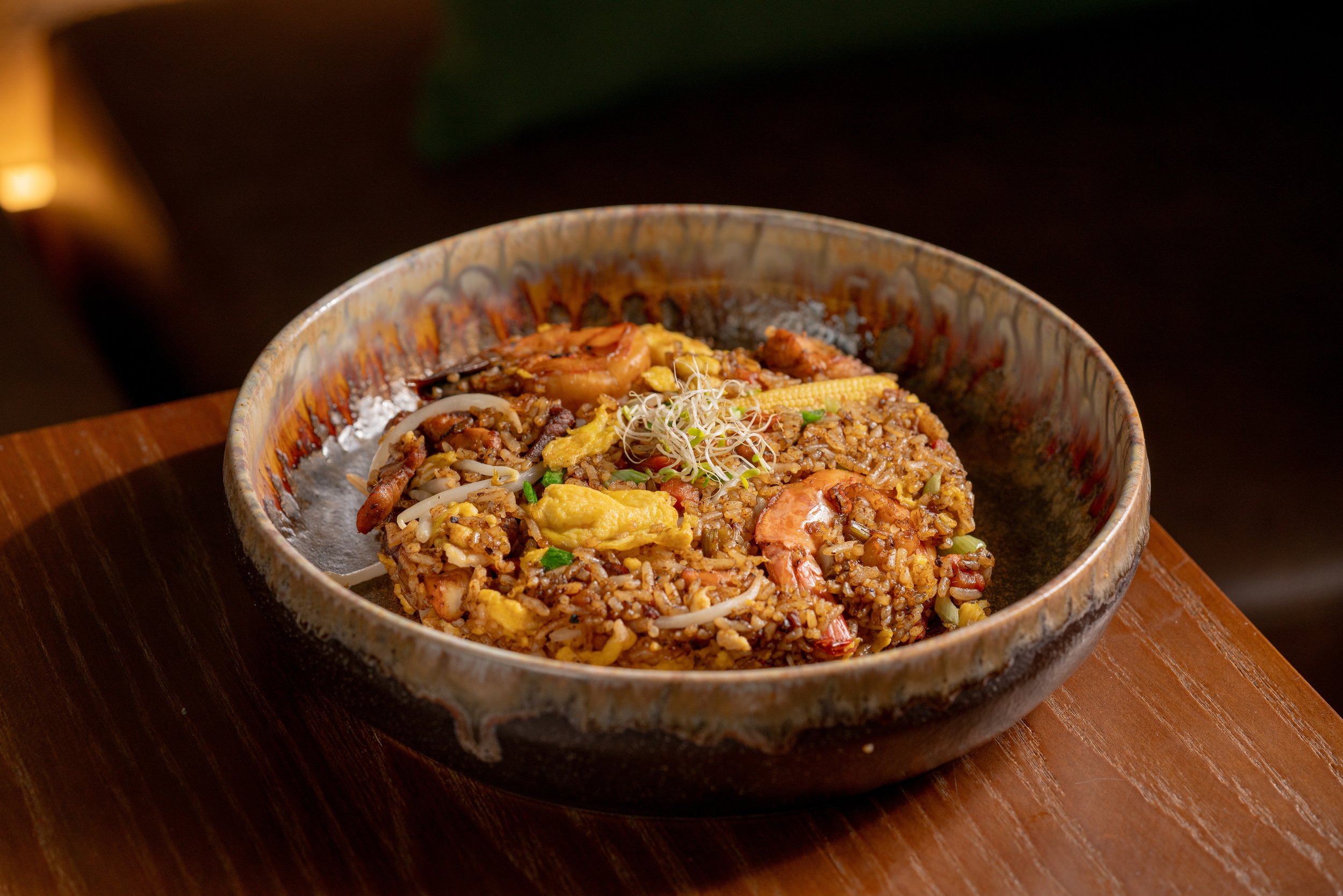 A ceramic bowl filled with fried rice, shrimp, scrambled egg, vegetables, and sprouts on top, placed on a wooden surface.