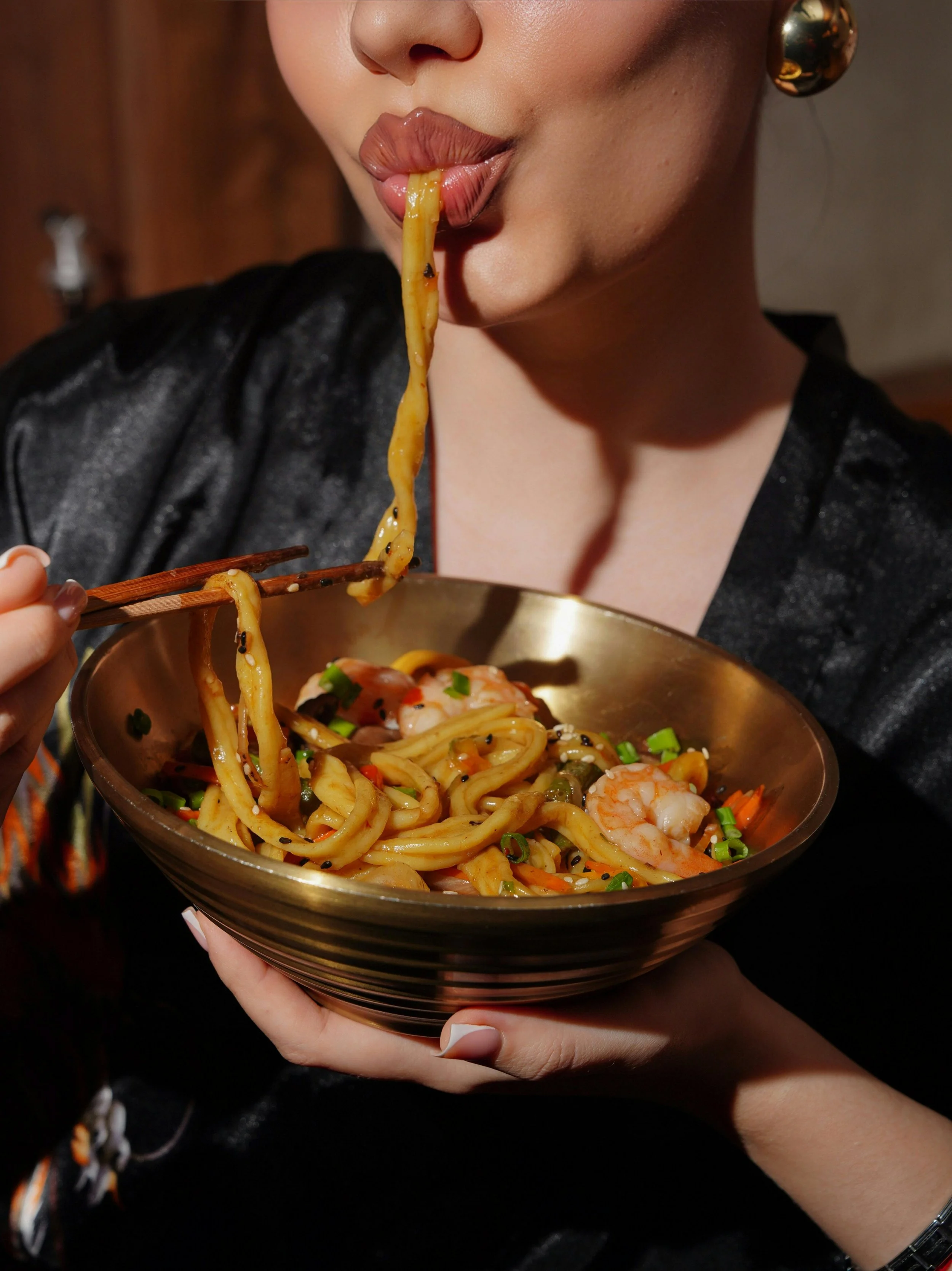 Person holding a bowl of Asian noodle dish with shrimp, vegetables, and sesame seeds, lifting noodles with chopsticks.