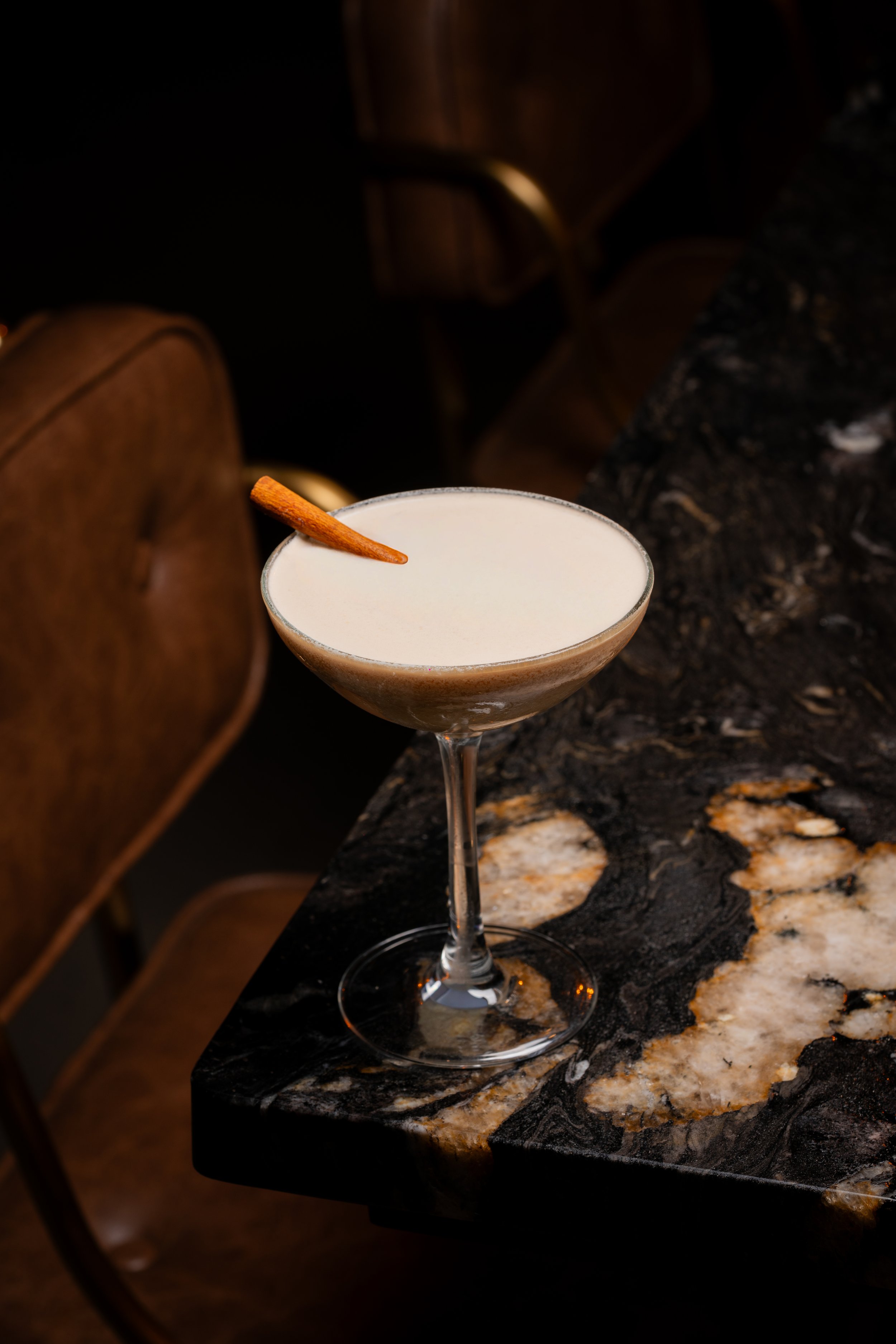 A cocktail glass with a light-colored drink, garnished with a cinnamon stick, placed on a dark marble counter.
