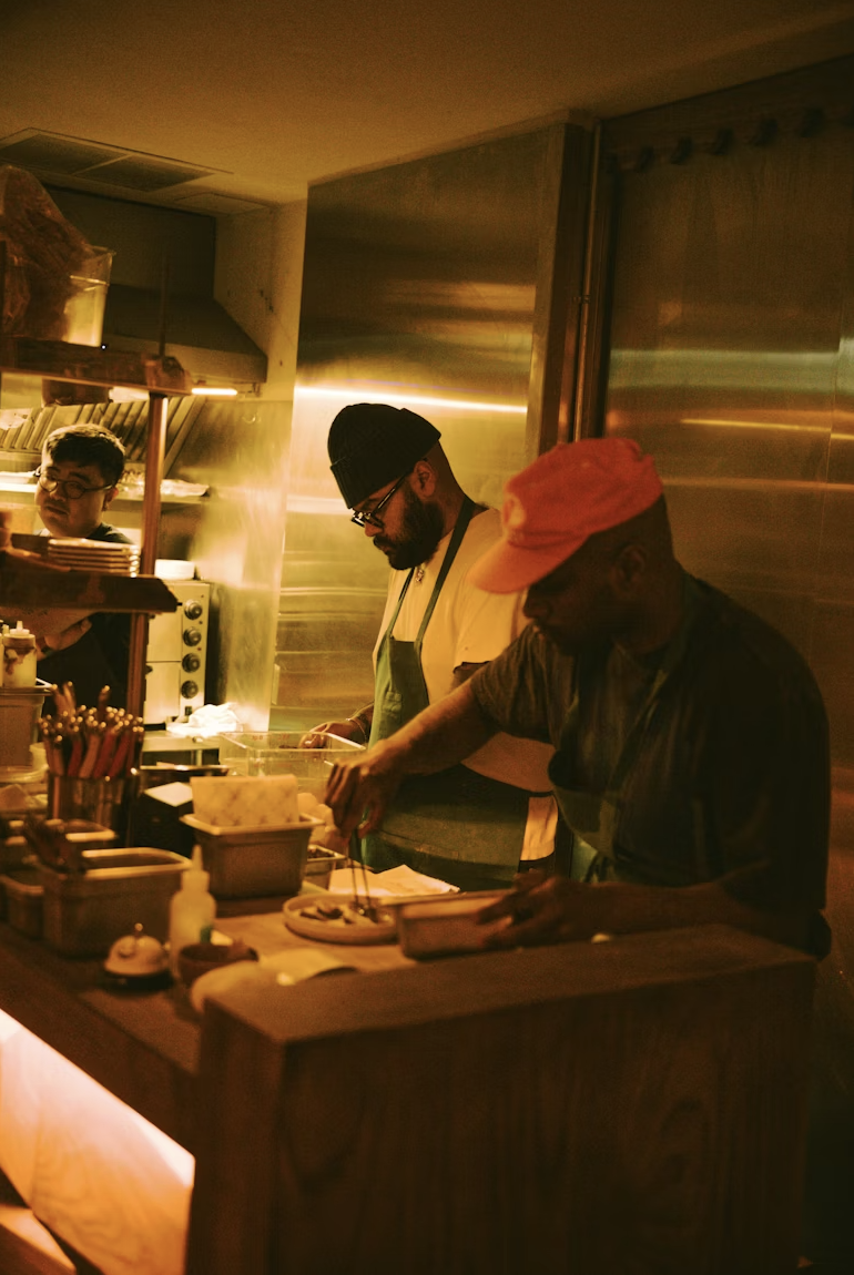 Three men working in a dimly lit kitchen, preparing food at a counter with various kitchen tools and ingredients.