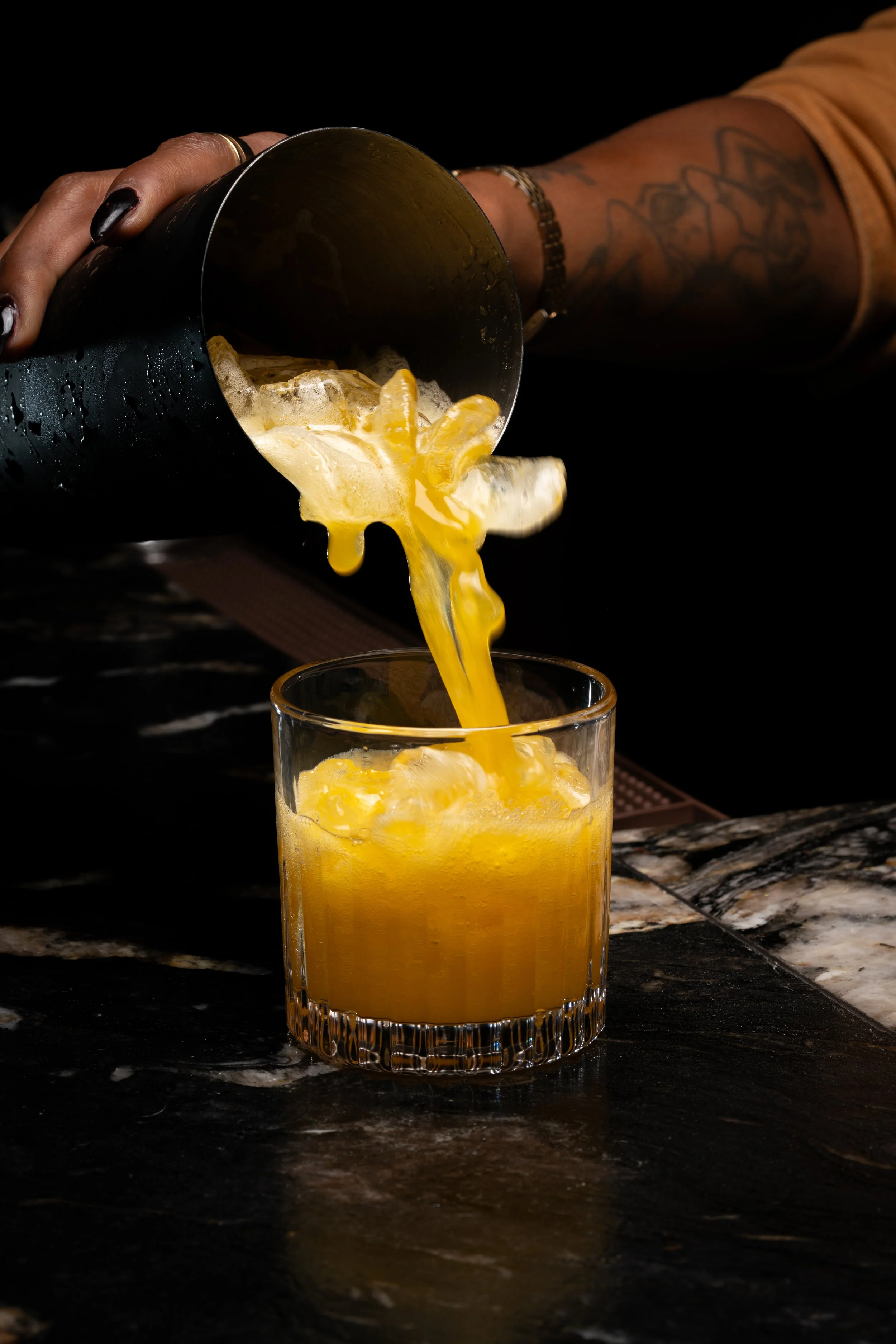 A person pouring a yellow-orange drink with ice cubes into a glass.
