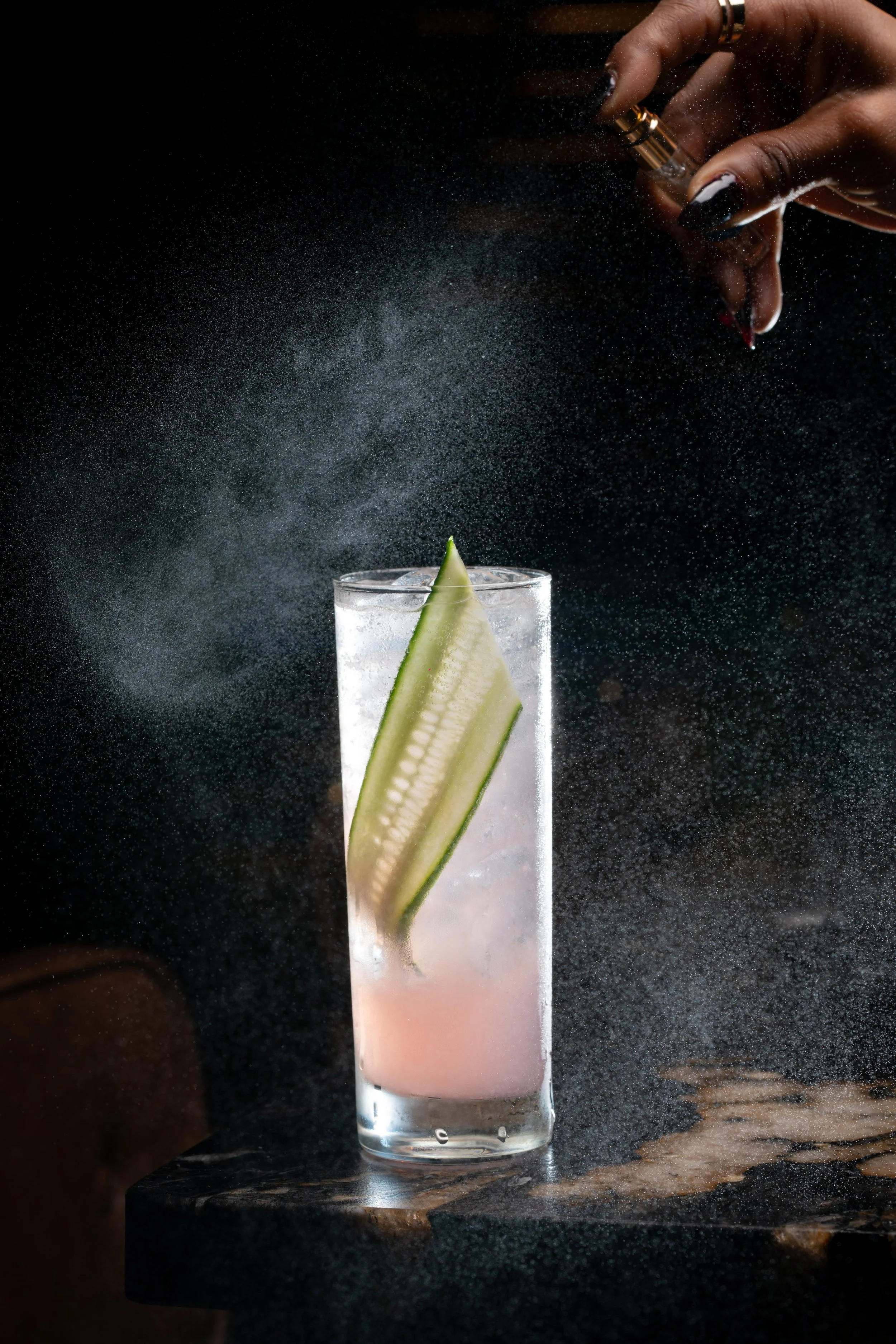 A tall glass of a pink layered drink with a slice of cucumber inside.