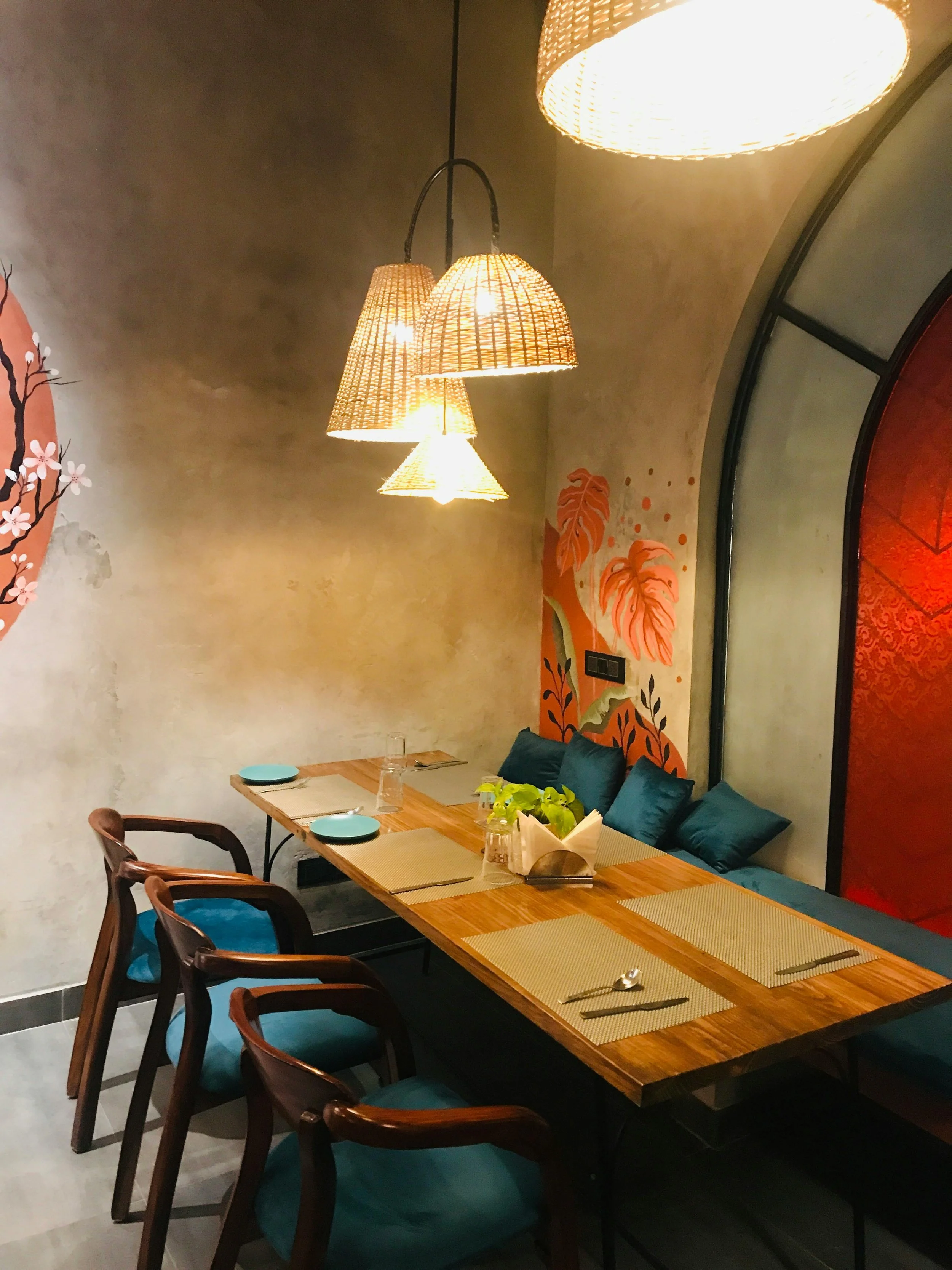 A dining room with a rectangular wooden table set for four, with placemats, utensils, and blue pillows along the wall. There are dark wooden chairs with blue cushions, and a decorative wall with orange and black painted leaves and flowers. Hanging above are four basket-like pendant lights providing warm lighting.
