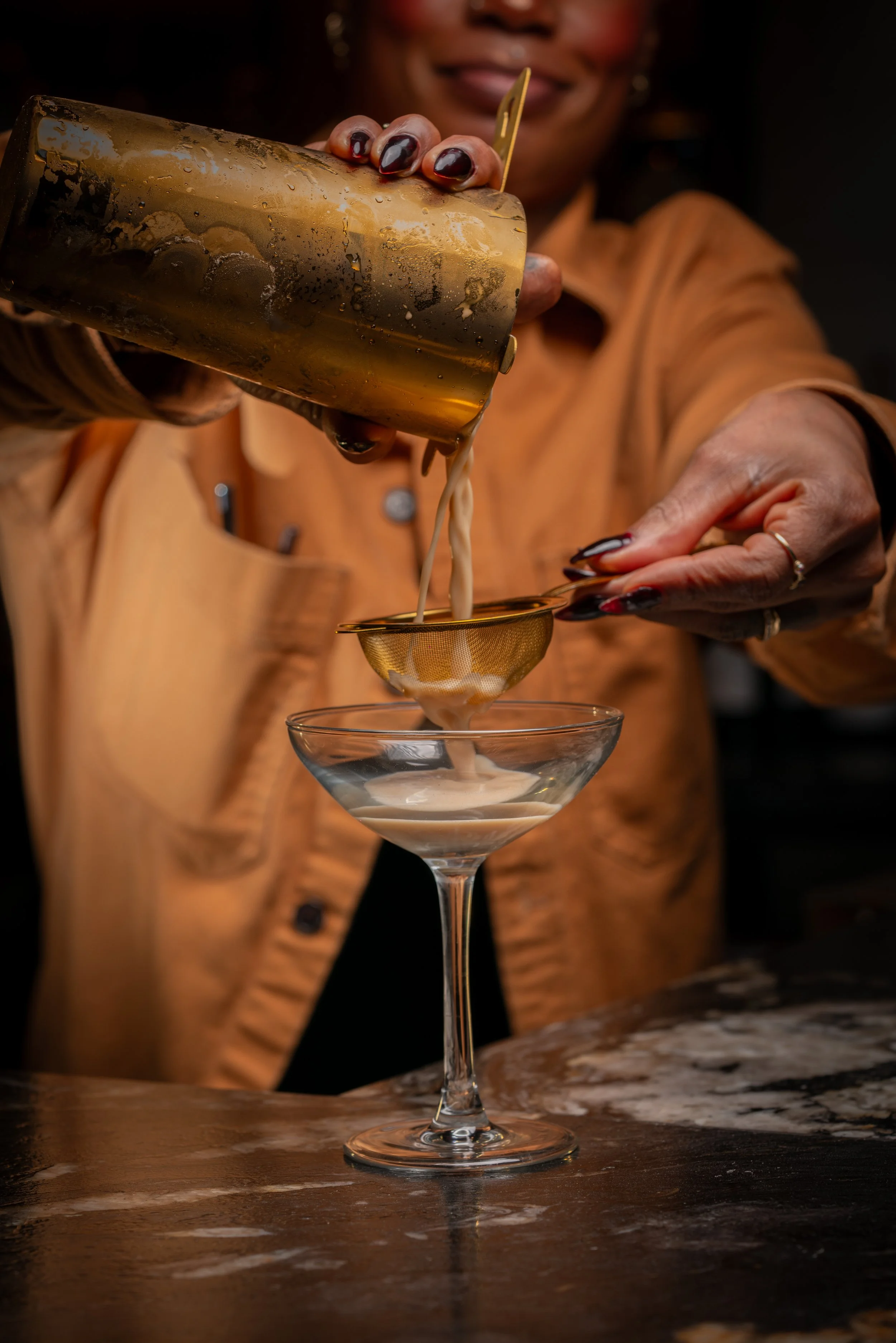 A woman pouring a creamy cocktail from a gold shaker into a martini glass, with a marble countertop in the foreground.