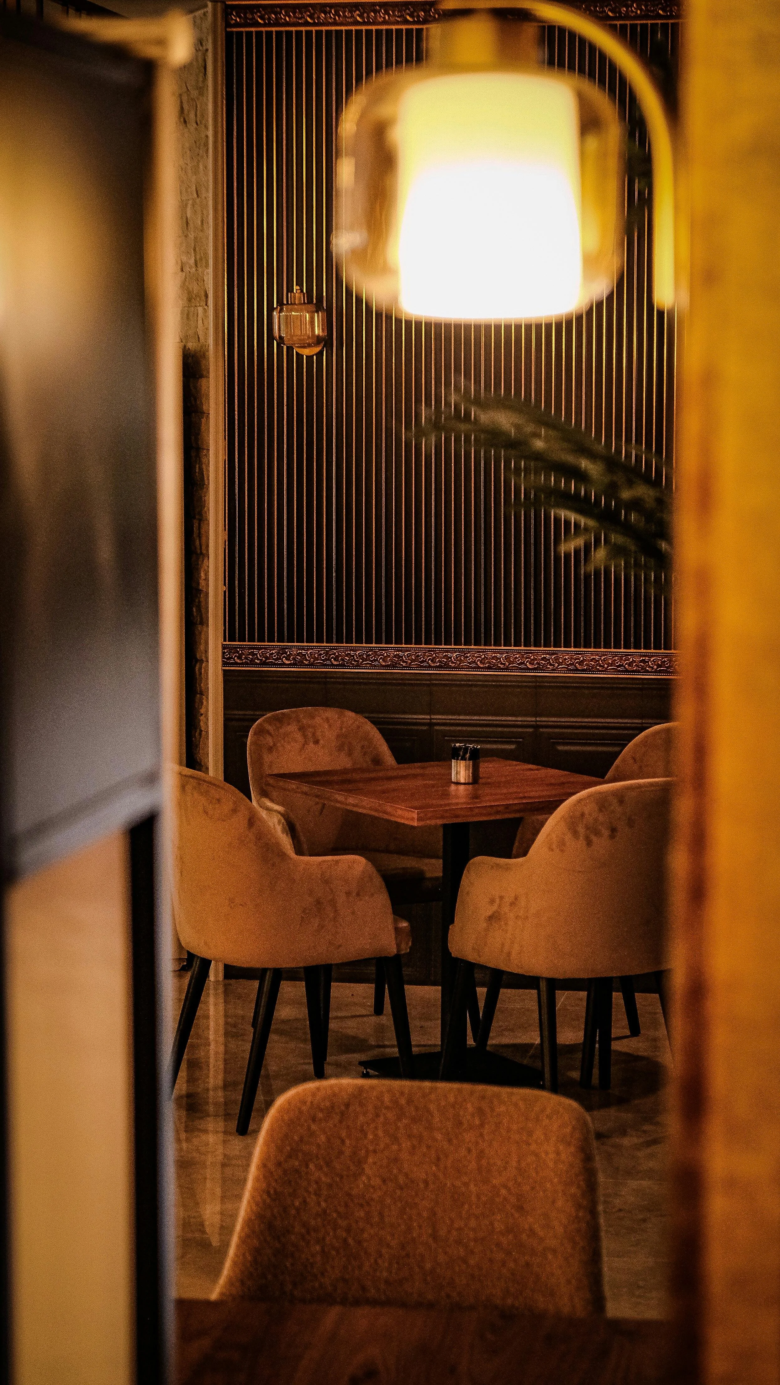 A cozy restaurant interior with a round wooden table, surrounded by plush, beige upholstered chairs. The background features a dark wall with vertical wooden slats, a decorative border, and a large hanging lampshade emitting warm light, creating an intimate atmosphere.
