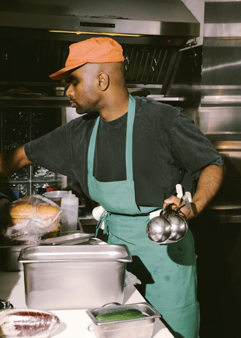 A man wearing an orange cap, black t-shirt, and a green apron working in a professional kitchen.