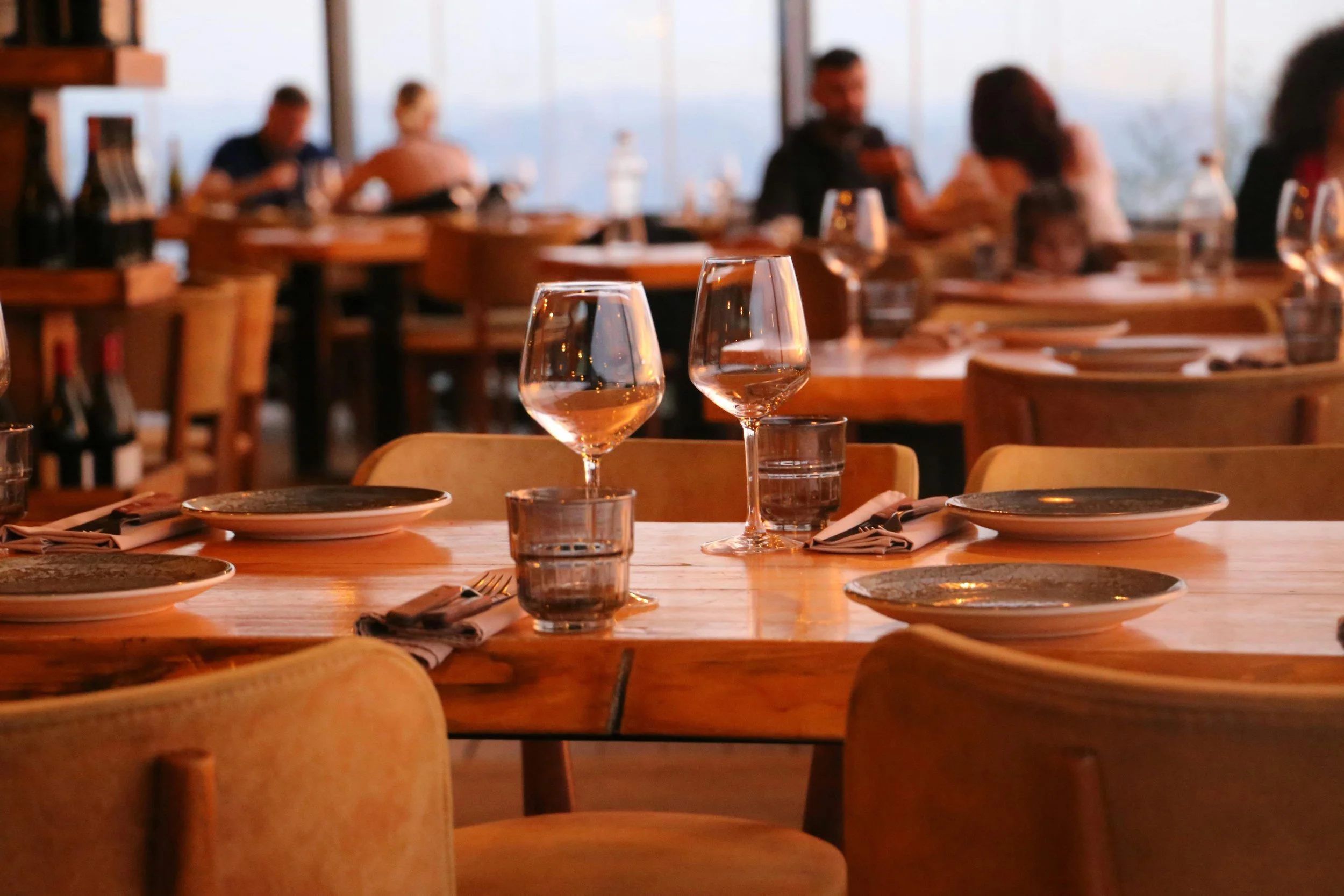 A dining table set with wine glasses, plates, and utensils inside a restaurant, with blurred people dining in the background.