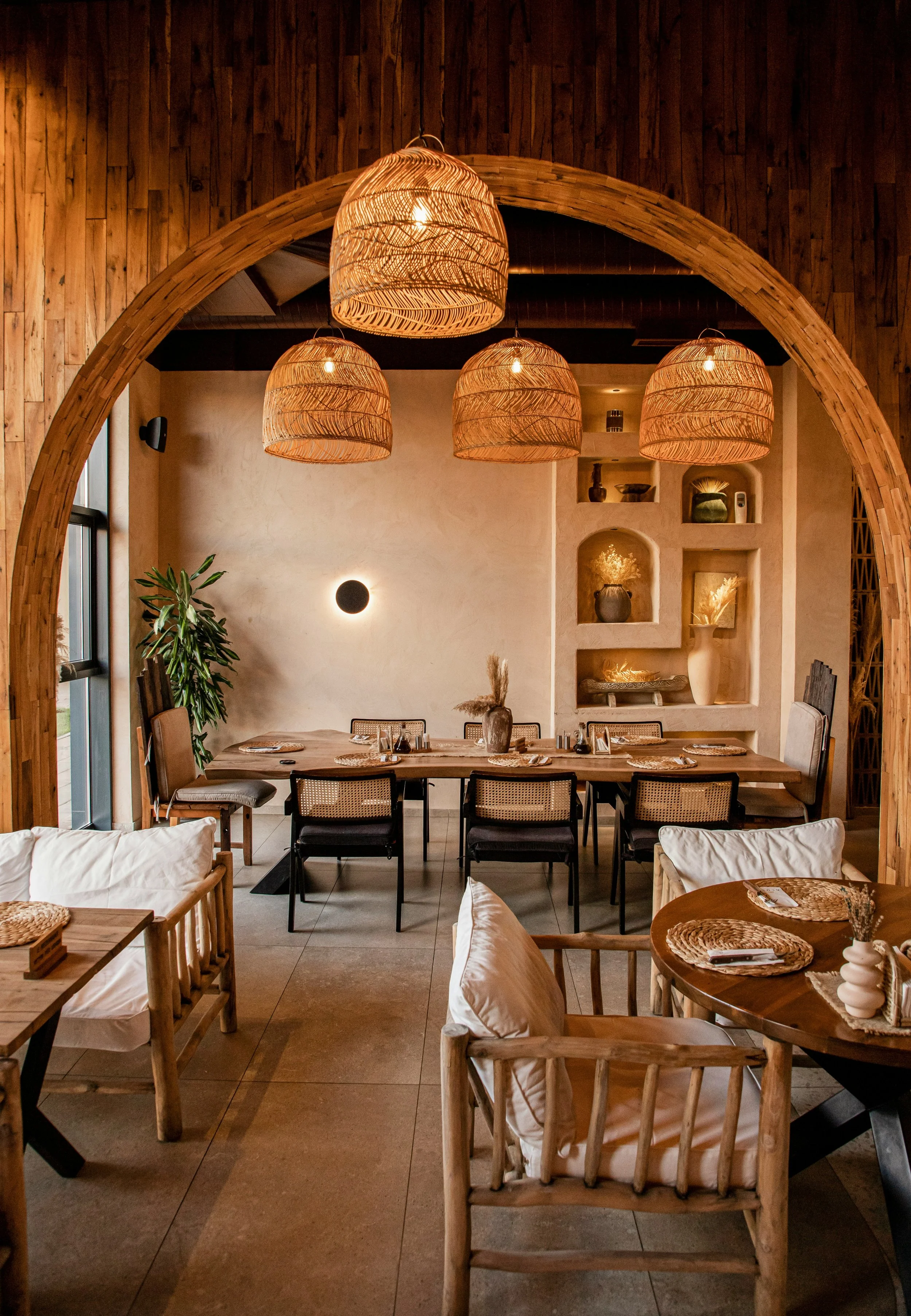 A cozy dining room decorated with wooden accents and hanging wicker lamps. The table is set with placemats, and there are chairs with white cushions around it. The room has natural light coming from a window on the left, and a built-in niche with decorative vases and sculptures on the back wall.