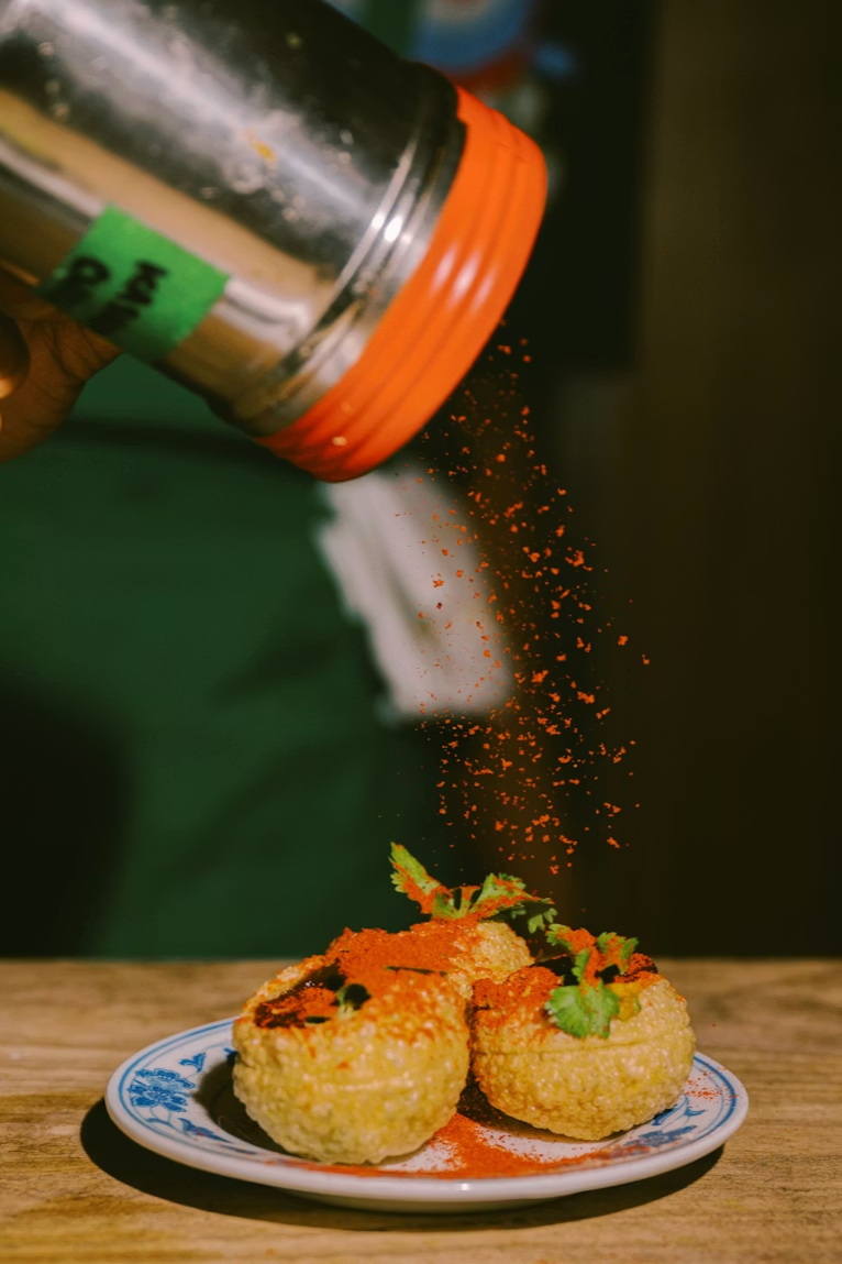 Deep-fried balls being sprinkled with orange seasoning over a decorative plate of food garnished with herbs.