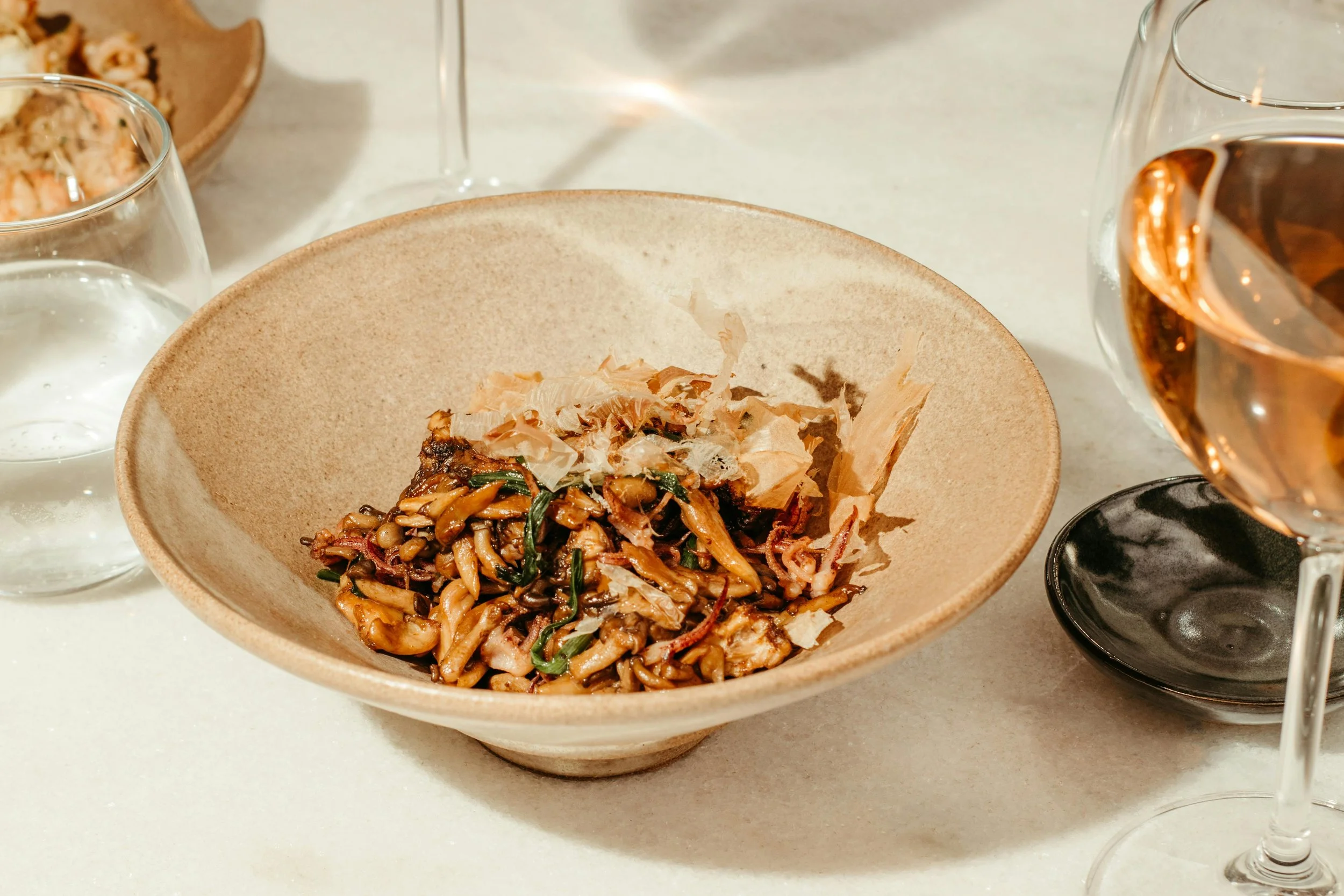 A bowl of stir-fried noodles with vegetables and bonito flakes, surrounded by glasses of water and wine on a white table.