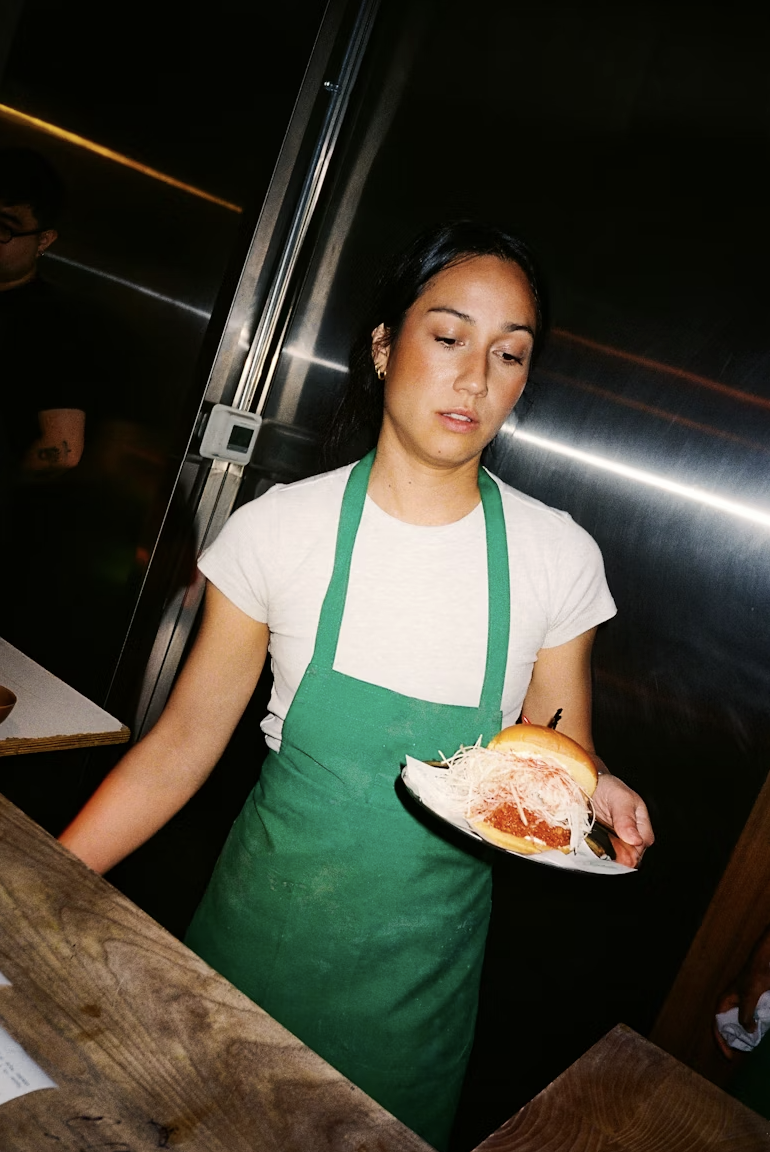 A woman in a white t-shirt and green apron holding a plate of food in a restaurant kitchen.