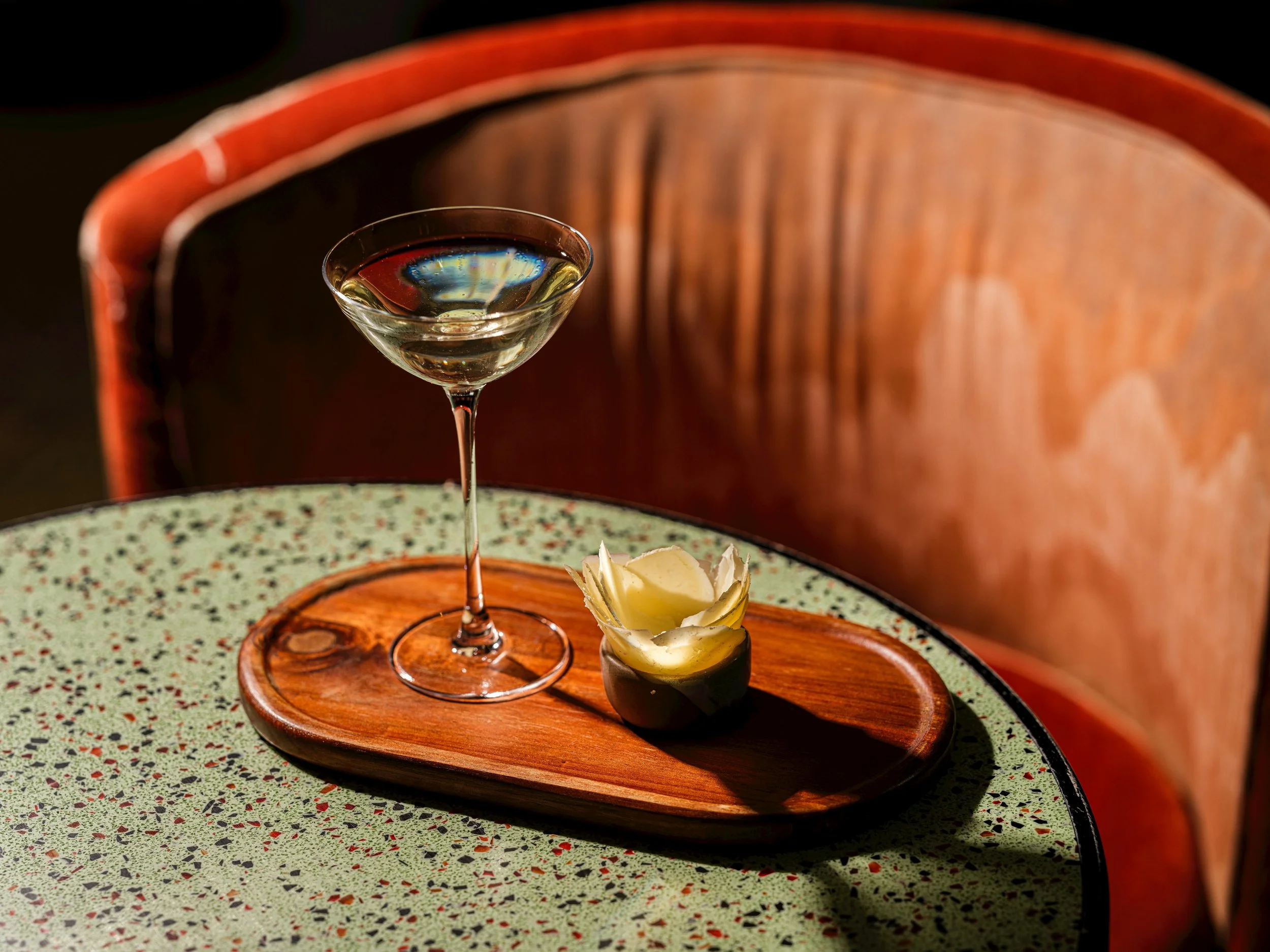 A glass of white wine on a wooden tray with cheese and a garnish, set on a speckled green and black table with a wooden chair in the background.
