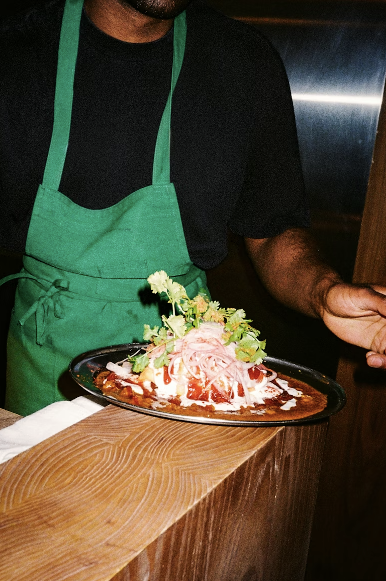 Person wearing a black shirt and green apron serving a dish of Mexican food topped with cilantro, onions, and sauce.