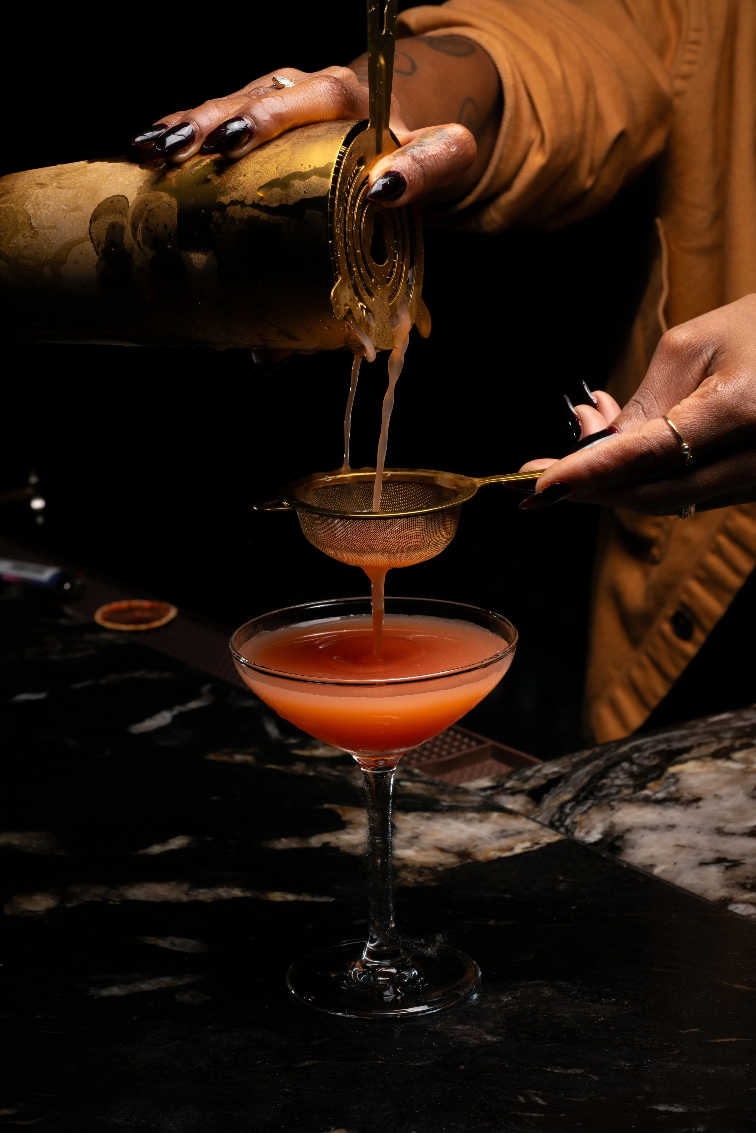 A person pouring a cocktail from a gold shaker into a glass with a gold strainer over a dark marble countertop.