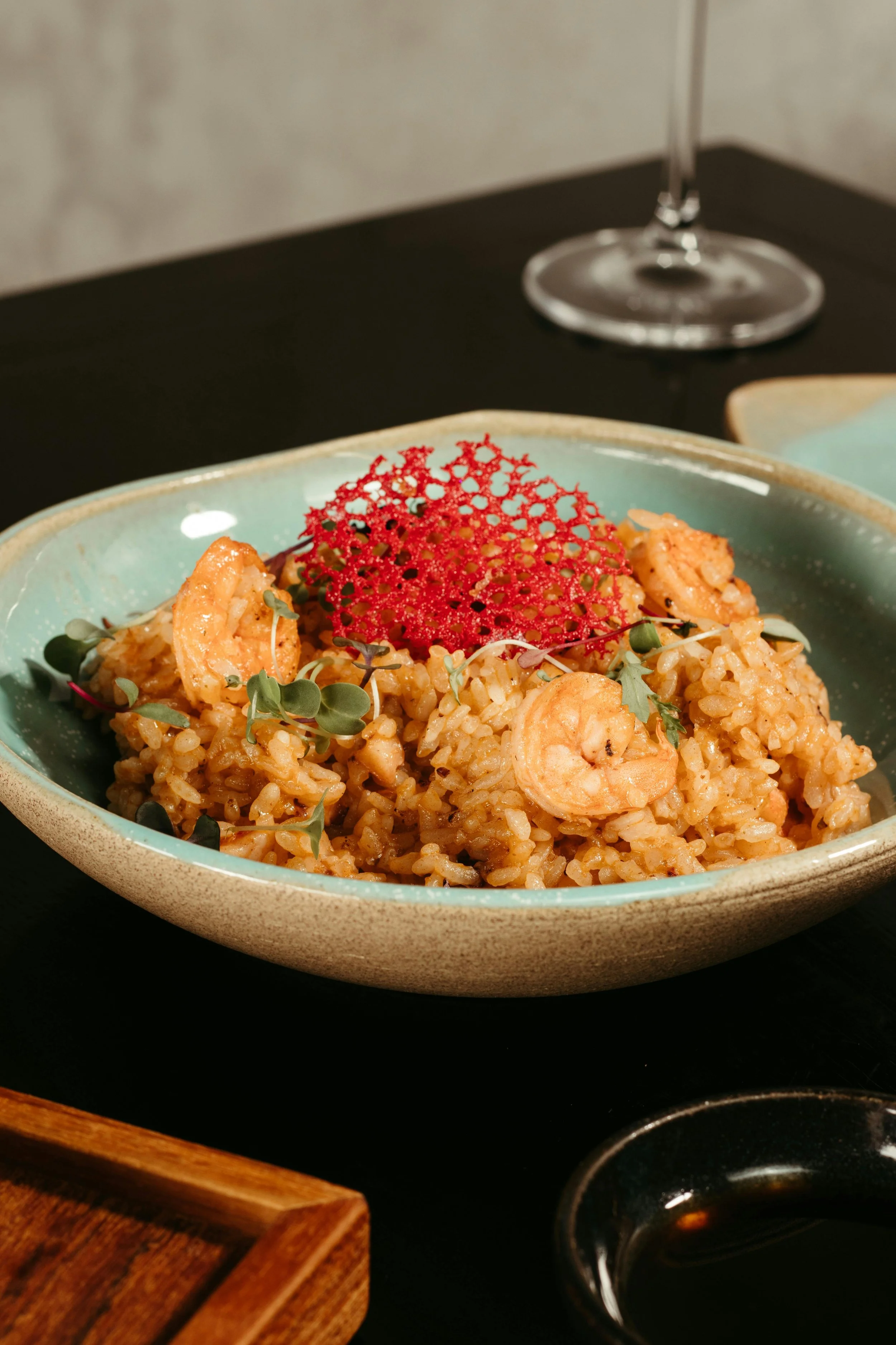 A close-up of a bowl of shrimp fried rice garnished with microgreens and a red crunchy garnish. In the background, there is a wine glass on a black table, and part of a wall and a wooden tray are visible.