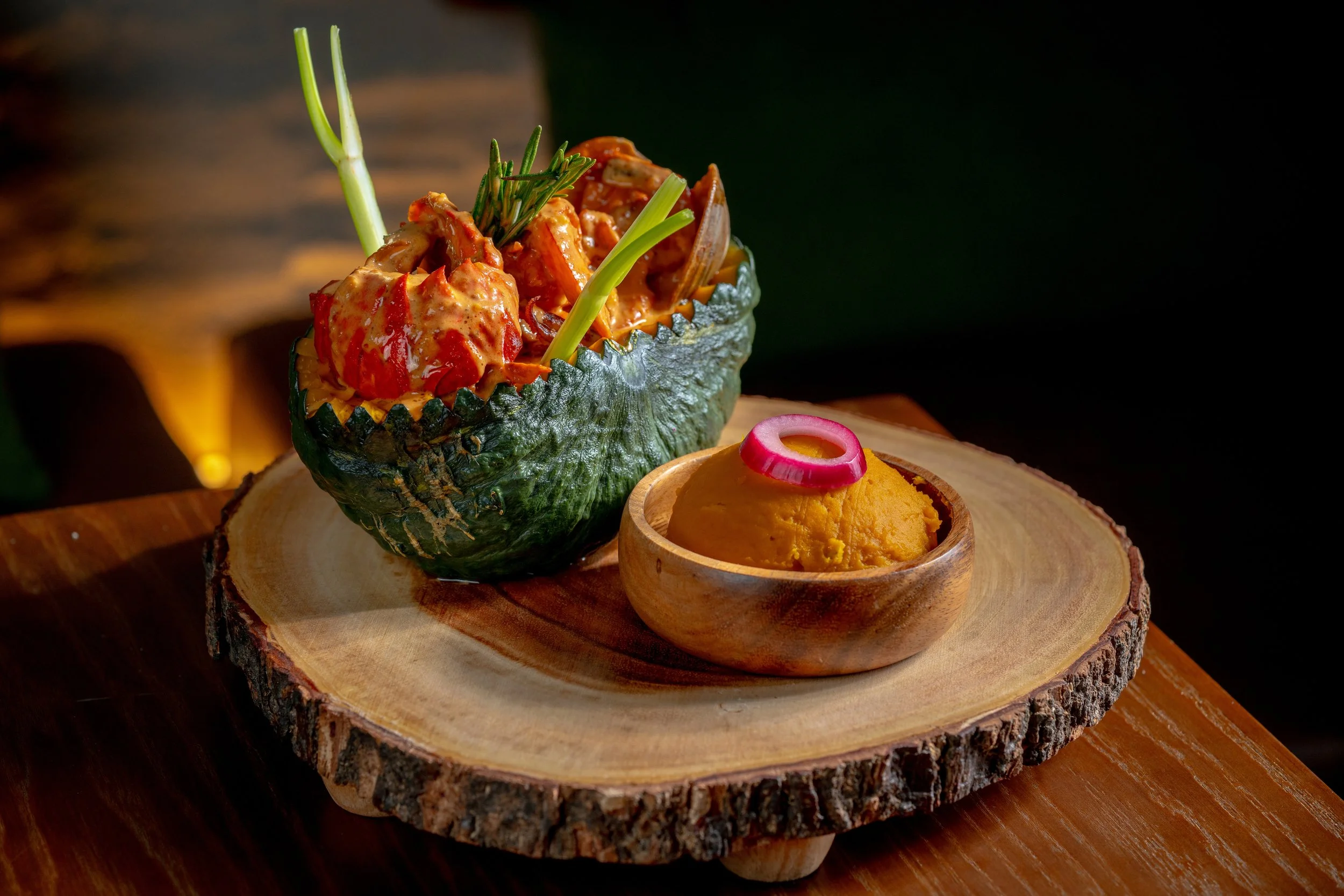 A carved-out green pumpkin filled with seafood, placed on a wooden slab with a small bowl of pumpkin purée.