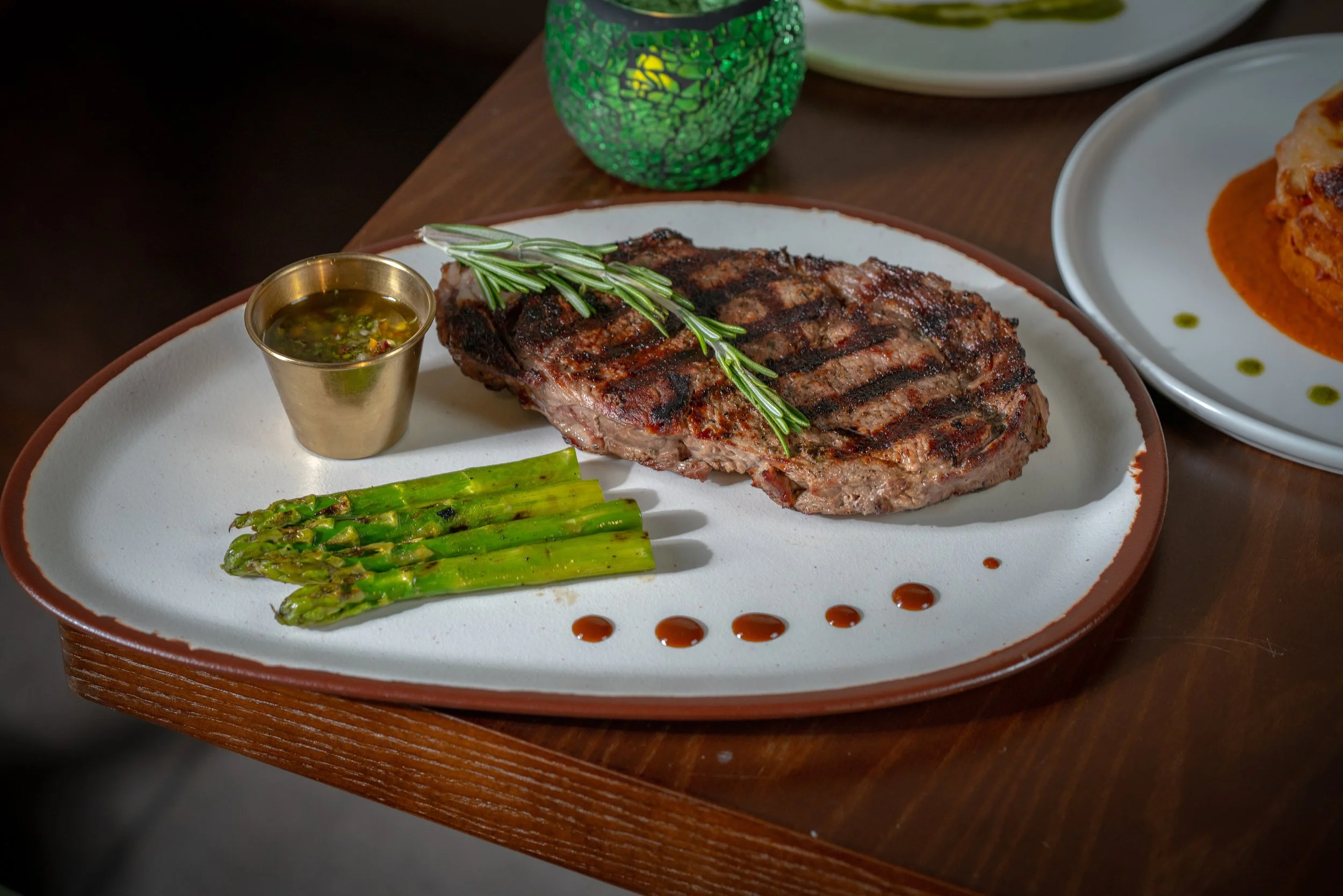 Grilled steak garnished with rosemary on a white plate, served with grilled asparagus and a small cup of sauce, with a green decorative candle in the background.