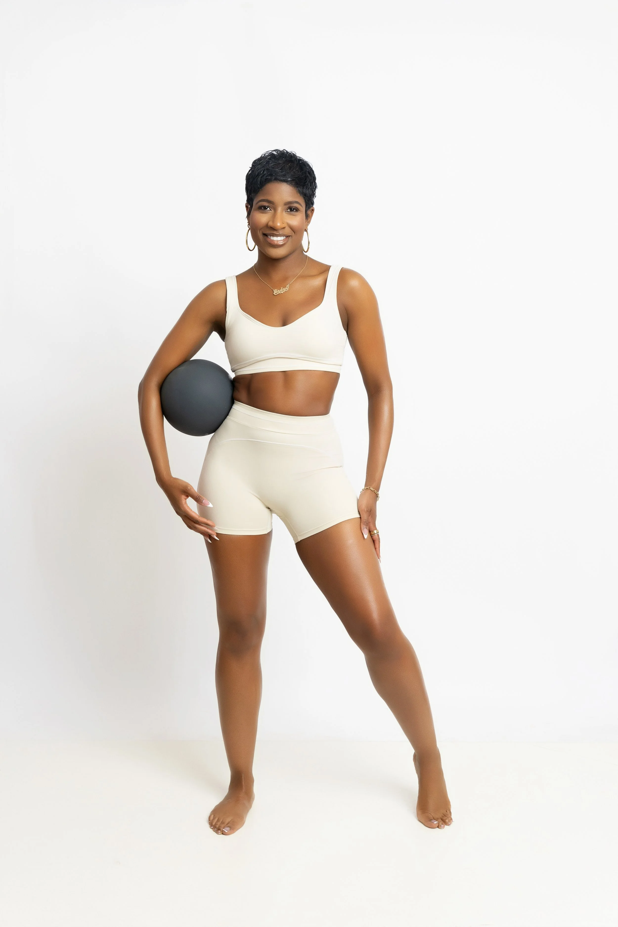 A woman standing barefoot against a plain white background, holding a black exercise ball under her right arm, wearing a cream-colored sports bra and matching shorts, with short curly black hair, gold hoop earrings, a gold necklace, and a gold bracelet.