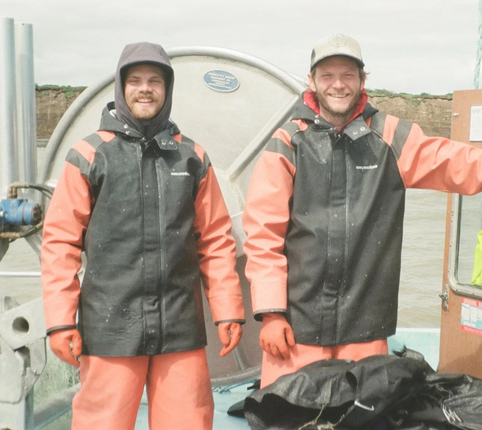 Two men in orange and black waterproof jackets and orange pants, smiling, standing in front of a white tank, outdoors on a cloudy day.