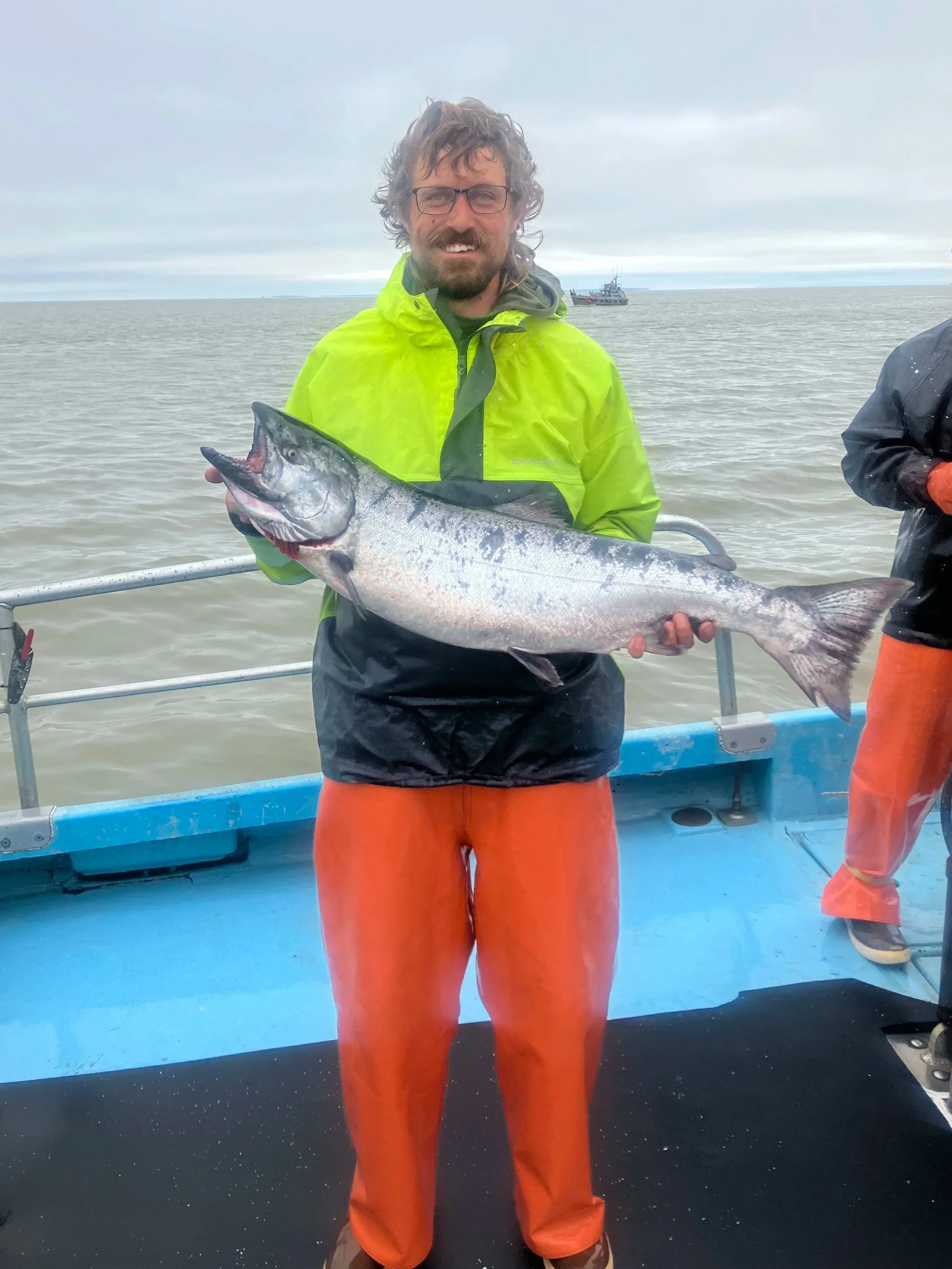 A person holding a large fish on a boat at sea, wearing an orange waterproof suit and a bright yellow rain jacket, with gray water and cloudy sky in the background.