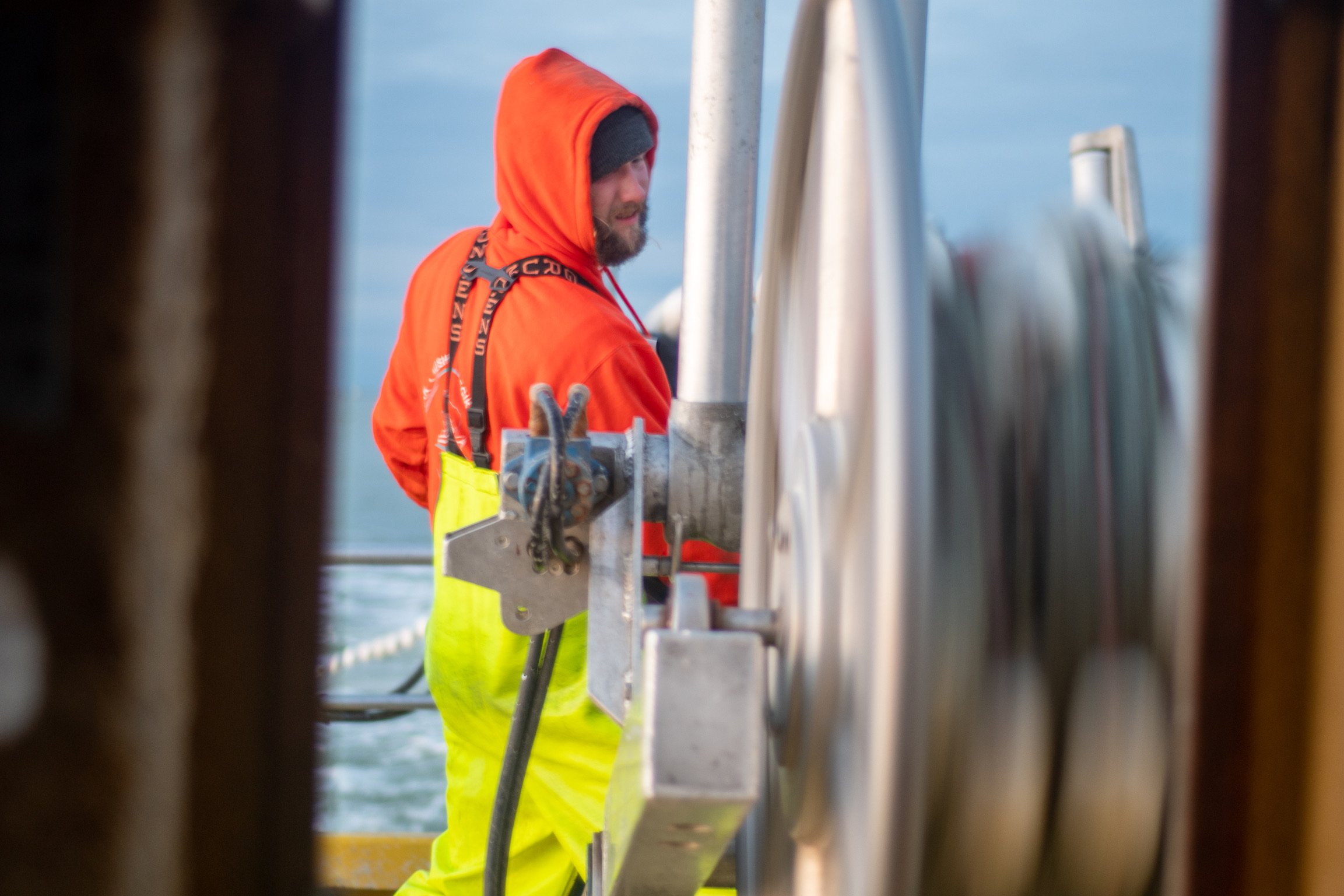 A person wearing a bright orange hoodie with the hood up, a gray beanie, and yellow safety pants, working on a boat or ship's rigging or maintenance, seen through the gaps between parts of the structure.