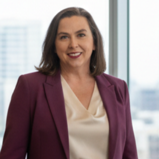 Portrait of a woman with shoulder-length brown hair, wearing a purple blazer and cream blouse, smiling in an office setting with large windows and cityscape background.