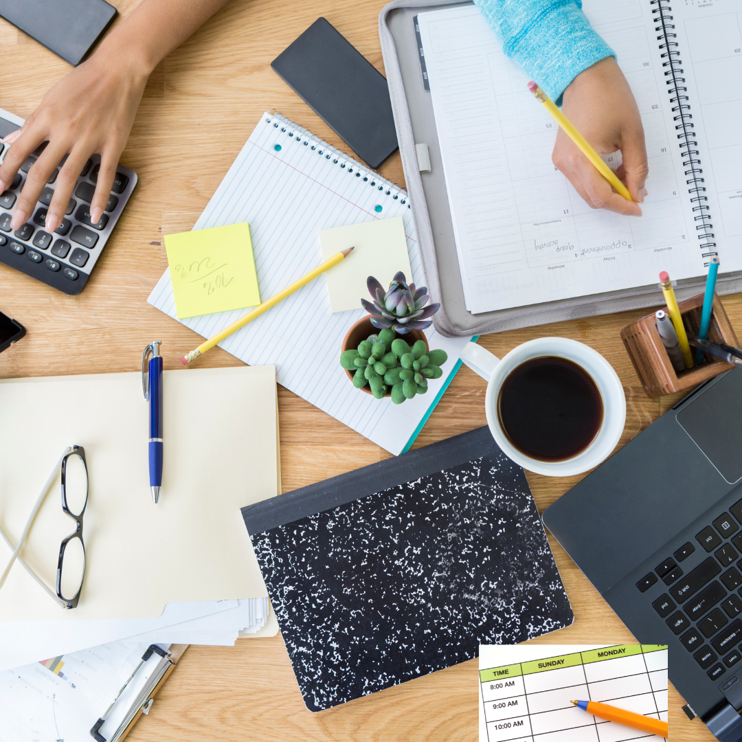 A cluttered wooden desk with a person's hands working, one on a calculator and the other writing in an organizer. The desk has a notebook, sticky notes, a cup of coffee, stationery supplies, a notebook with a black and white speckled cover, a pair of eyeglasses, a small potted succulent, and a printed weekly schedule.
