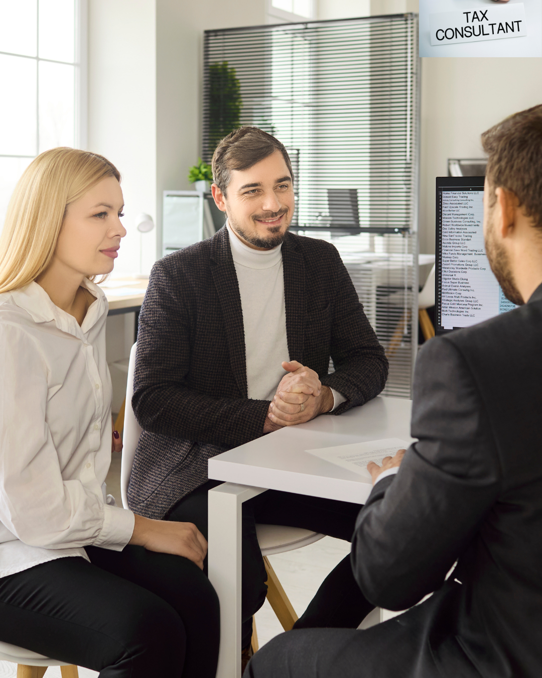 A man and woman sitting across from a man in a suit during a consultation in an office, with a sign reading 'Tax Consultant' hanging from the ceiling.