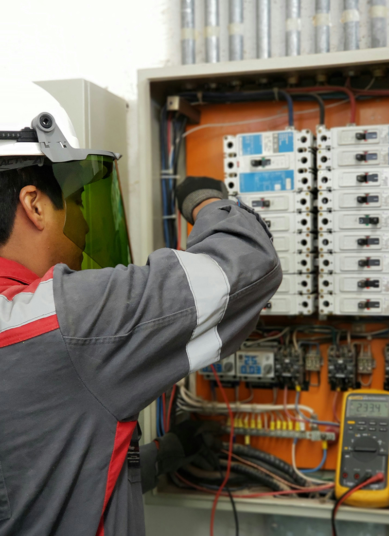 An electrician working on an electrical panel with circuit breakers and wiring, wearing safety gear including a helmet with a visor.