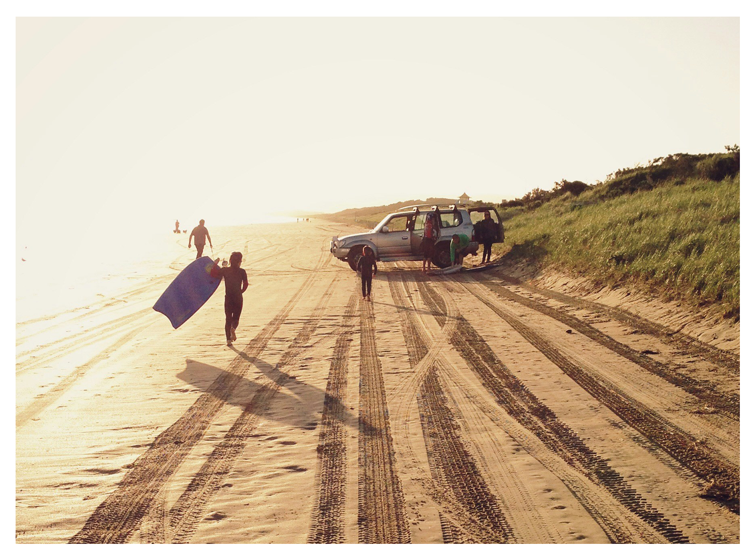 People on a beach with a vehicle parked on the sand and a person holding a surfboard, early morning or late afternoon sunlight casting long shadows.