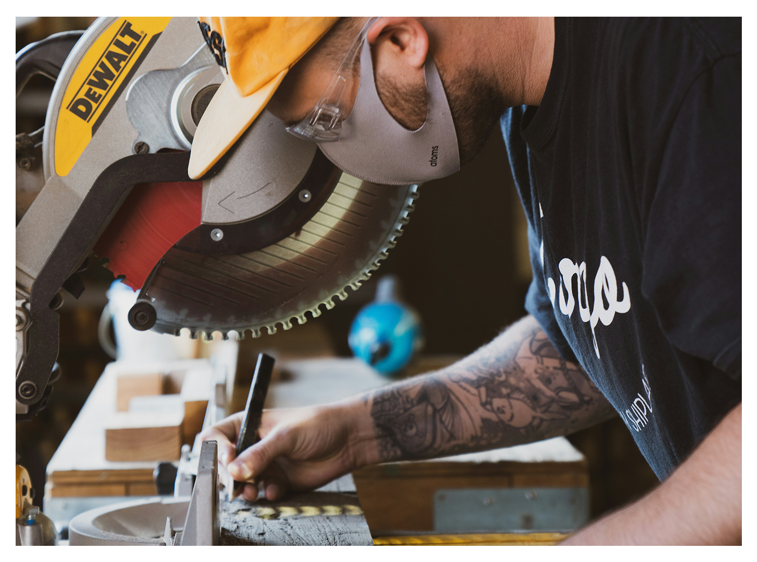A man working with a DeWalt miter saw, wearing safety glasses and a dust mask, cutting a piece of wood in a workshop.