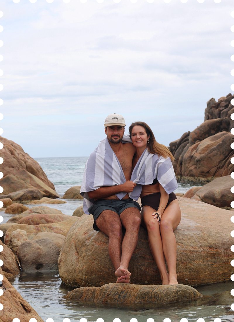 A couple sitting on a large rock at the beach, wrapped in striped towels, with rocky formations and the ocean in the background.