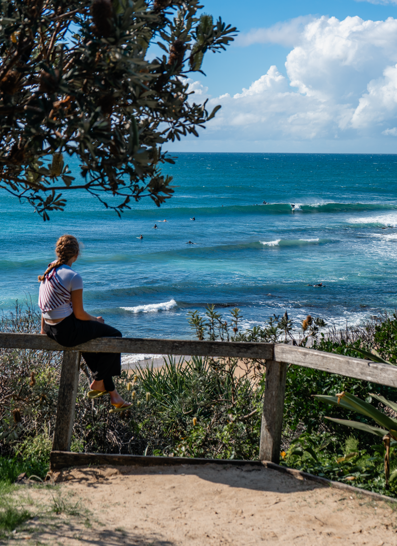 A girl with braids wearing a striped shirt and dark pants sitting on a wooden railing overlooking the ocean with surfers riding waves.