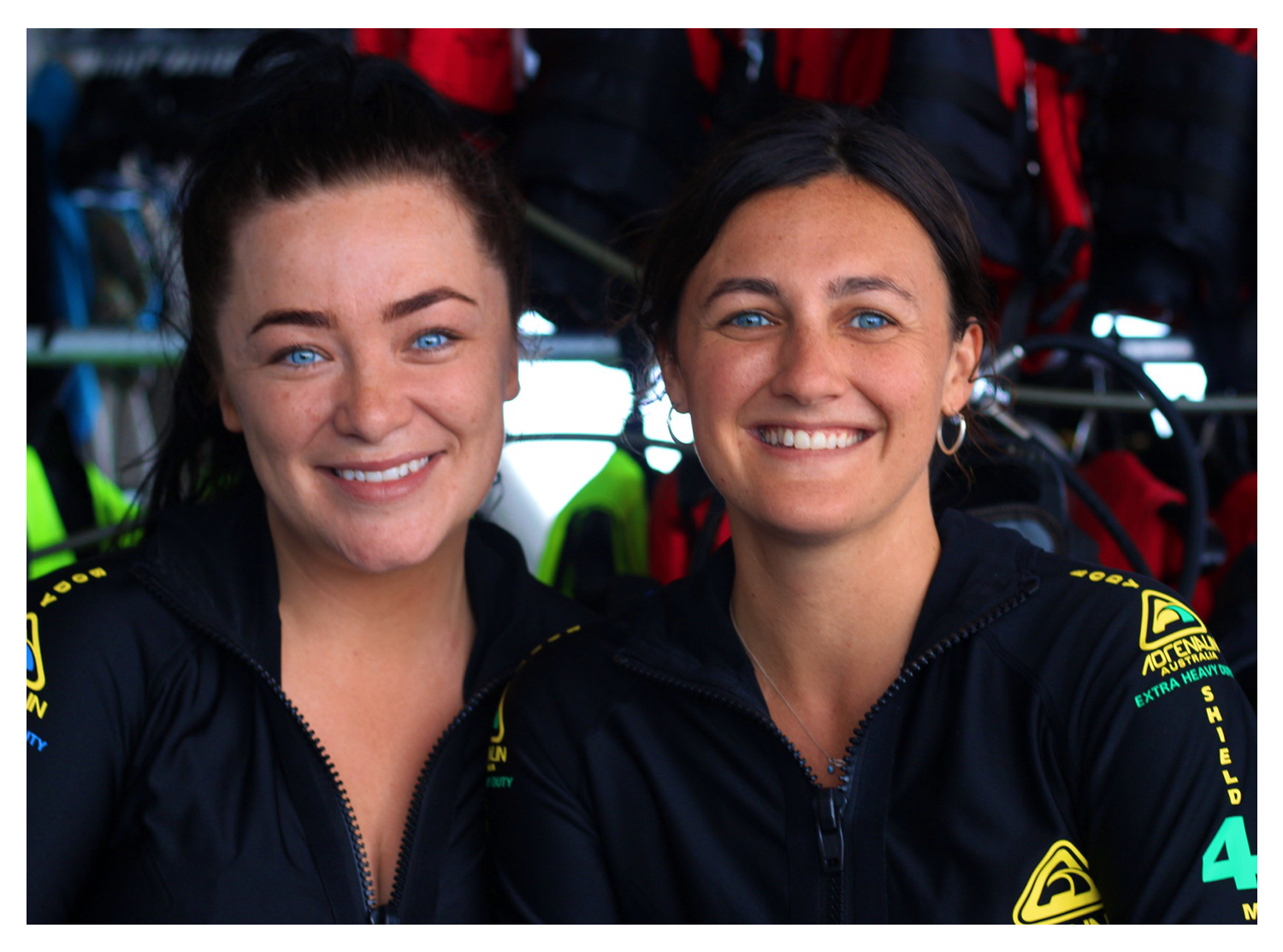 Two women with blue eyes smiling, wearing black jackets with yellow and green patches, possibly gear for outdoor activity or sports, against a background of backpacks and gear.