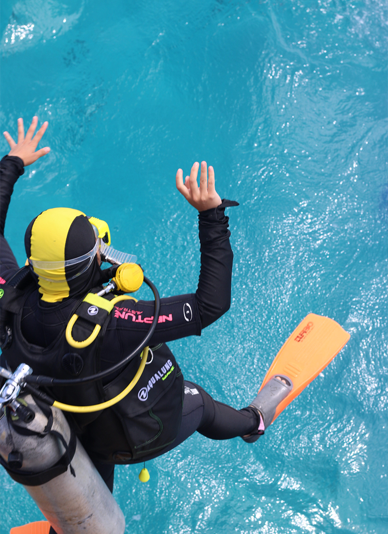 A person in scuba diving gear jumping into the water with fins on, surrounded by blue water.